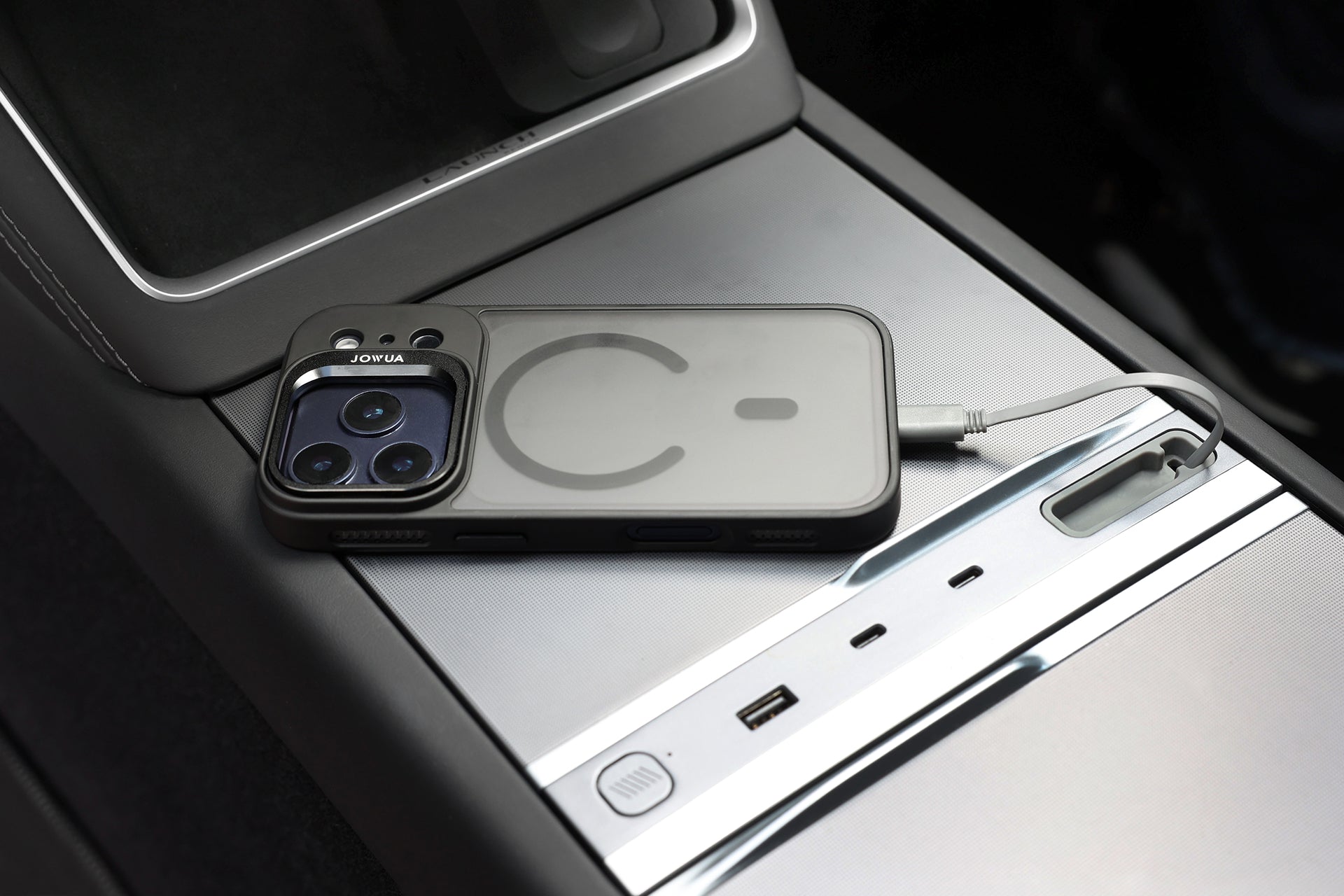 A JOWUA iPhone Case for Tesla Car Mount with MagSafe + Phone Tether Tab Set (iPhone 17 Series) is charging via USB-C on a car's center console, next to two USB ports and a small storage compartment.