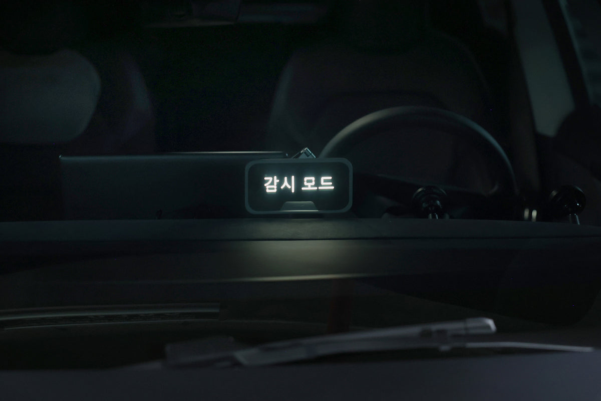 A digital sign with Korean text “감시 모드” (meaning “Surveillance Mode”) is displayed on a car dashboard, viewed through the windshield with the steering wheel and car interior visible. The scene subtly highlights modern features like the JOWUA MagSafe LED Light with Light Stencil Set, seamlessly integrated into the sleek design to provide convenient, customizable illumination for car interiors.
