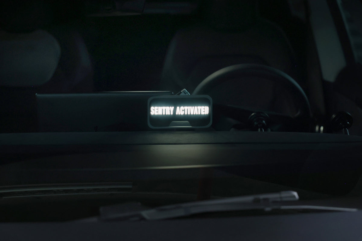A close-up of a car dashboard highlights a glowing MagSafe LED Light with Light Stencil Set by JOWUA displaying “Sentry Activated,” signaling the security system is engaged. The scene also showcases the sleek steering wheel, while beneath it, a Tesla wireless charging pad stands ready to conveniently power your devices. A windshield wiper can be seen as well.