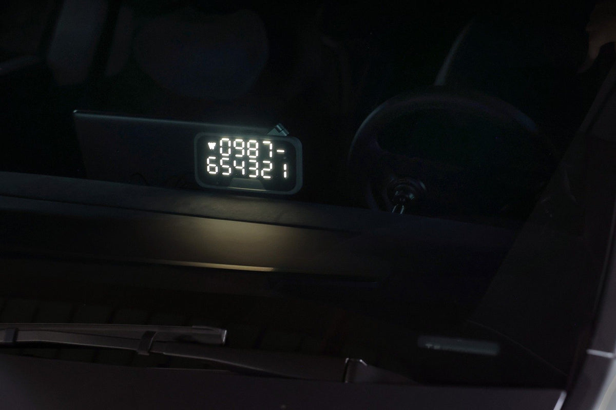 A digital display shows the numbers 0987 and 654321, seen through a car windshield with part of the dashboard and steering wheel visible in low light. On the console, a JOWUA MagSafe LED Light with Light Stencil Set adds subtle illumination, emphasizing seamless wireless charging and customizable lighting effects for your devices even on nighttime drives.