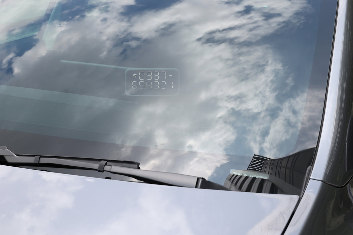 A car windshield reflects a cloudy sky. Inside the windshield, a digital display shows numbers and letters—likely a heads-up display—while a wiper rests at the bottom. On the dashboard, the JOWUA MagSafe LED Light with Light Stencil Set casts a subtle glow beside a Tesla wireless charging pad, blending modern convenience with sleek design.