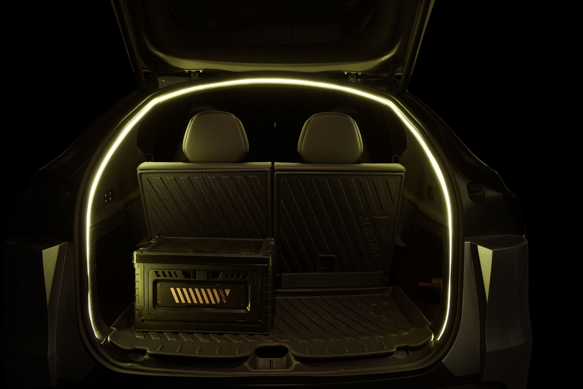 The image shows the open trunk of a car illuminated by yellow ambient LED lighting around the edge. Inside are two black seats, a folding crate, protective mats, and a JOWUA Tesla Model Y L (2026+) Trunk Storage Bins + Ambient Light Bundle trunk organizer, all set against a dark background.