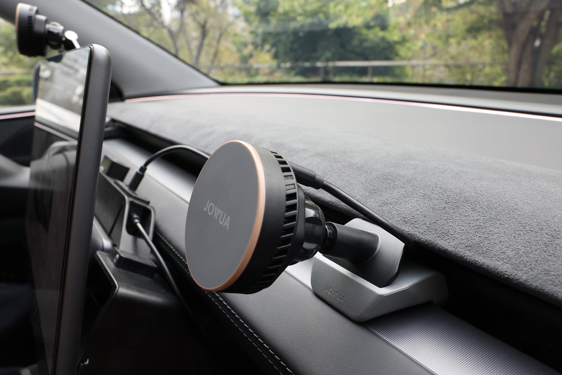 A close-up of a car dashboard shows the JOWUA Active Cooling MagSafe Car Mount for Air Vents (Model 3 Highland & Model Y Juniper), a tablet on a stand, and trees visible through the windshield.