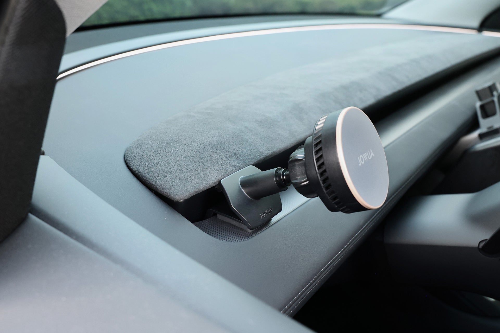 A close-up of a car dashboard shows a JOWUA Active Cooling MagSafe Car Mount for Air Vents (Model 3 Highland & Model Y Juniper) near the windshield, with part of the driver’s seat and steering wheel visible.