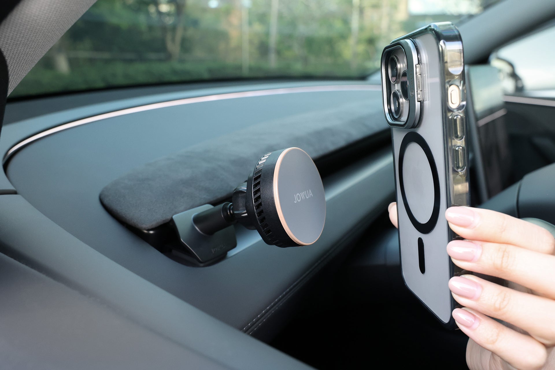 A person holds a smartphone with a ring-shaped MagSafe case near the JOWUA Active Cooling MagSafe Car Mount for Air Vents (Model 3 Highland & Model Y Juniper), which supports MagSafe wireless charging and is attached to the car dashboard.