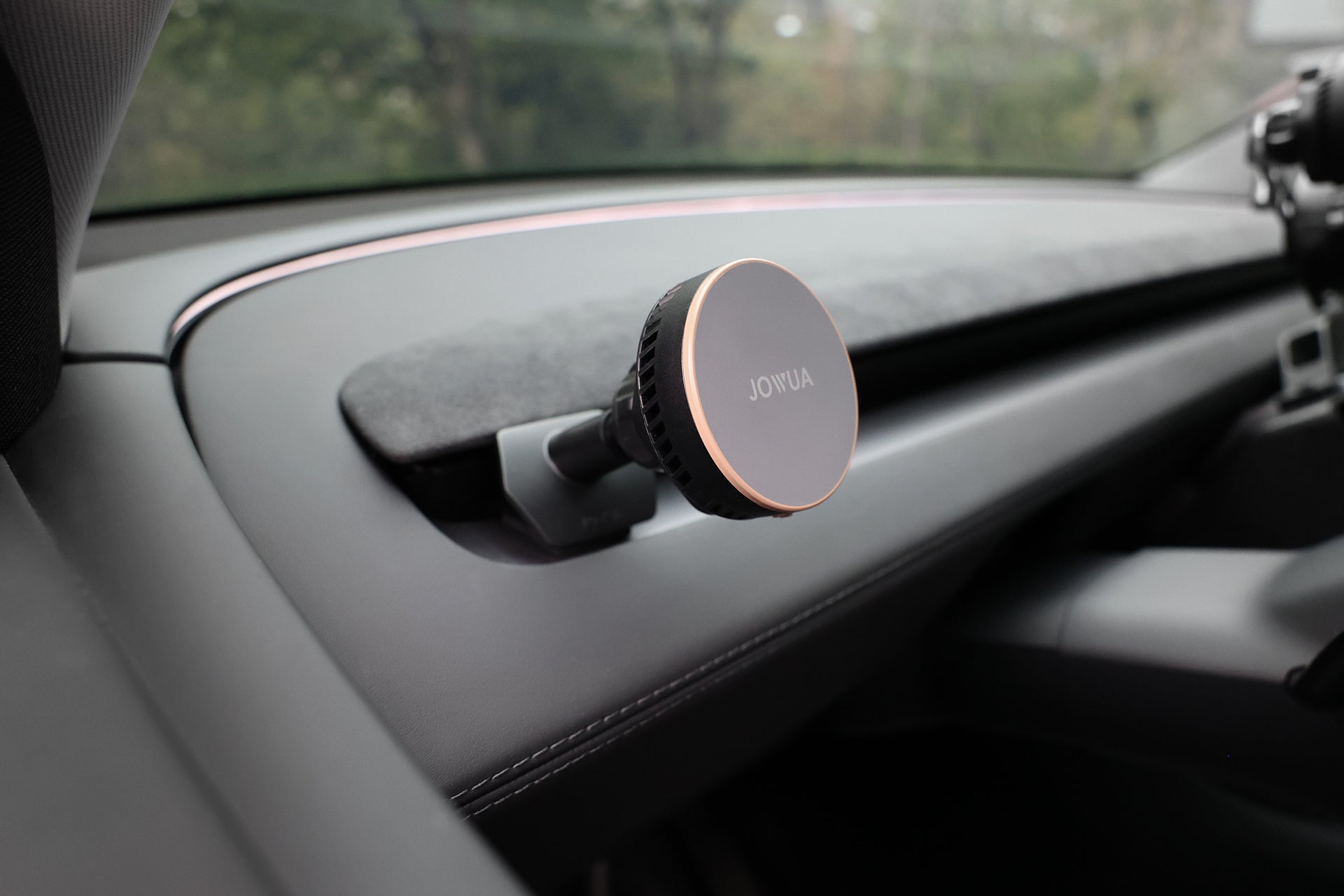 A close-up of a JOWUA Active Cooling MagSafe Car Mount for Air Vents (Model 3 Highland & Model Y Juniper) on a car dashboard, with blurred trees visible through the window.