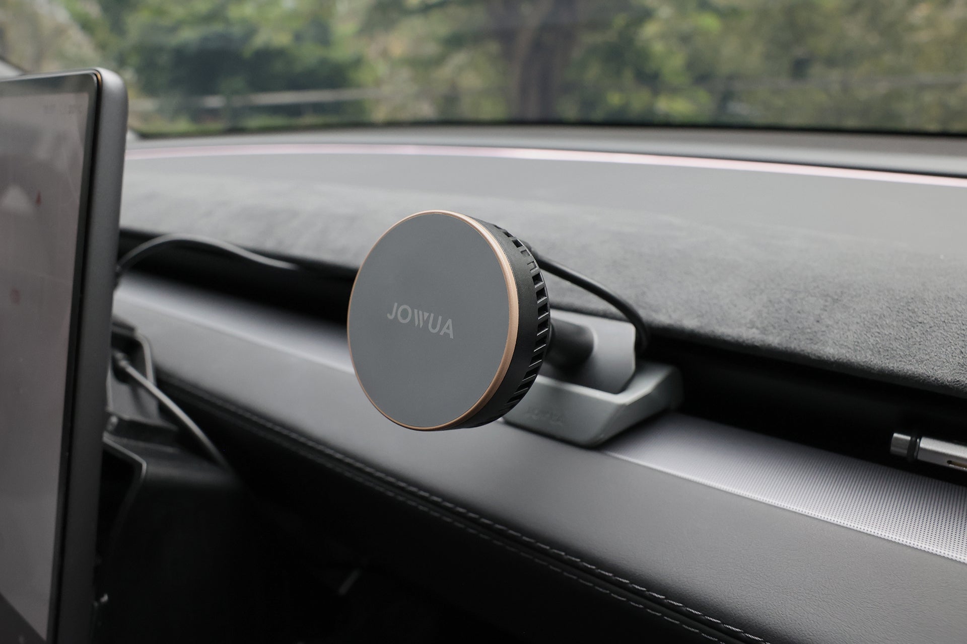 A round, black JOWUA Active Cooling MagSafe Car Mount for Air Vents (Model 3 Highland & Model Y Juniper) is attached to a car dashboard, with trees and greenery visible through the windshield in the background.