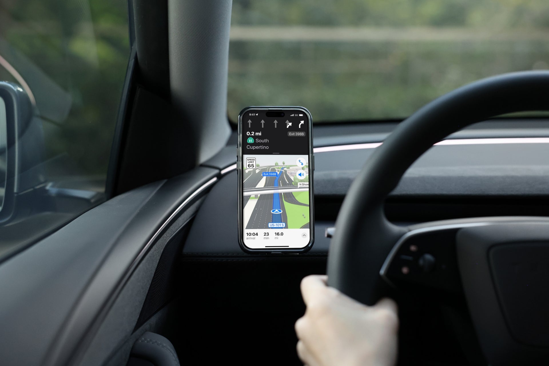 A person drives with a JOWUA Active Cooling MagSafe Car Mount for Air Vents (Model 3 Highland & Model Y Juniper) on the dashboard, holding their smartphone for GPS navigation, one hand on the wheel, with trees visible outside.
