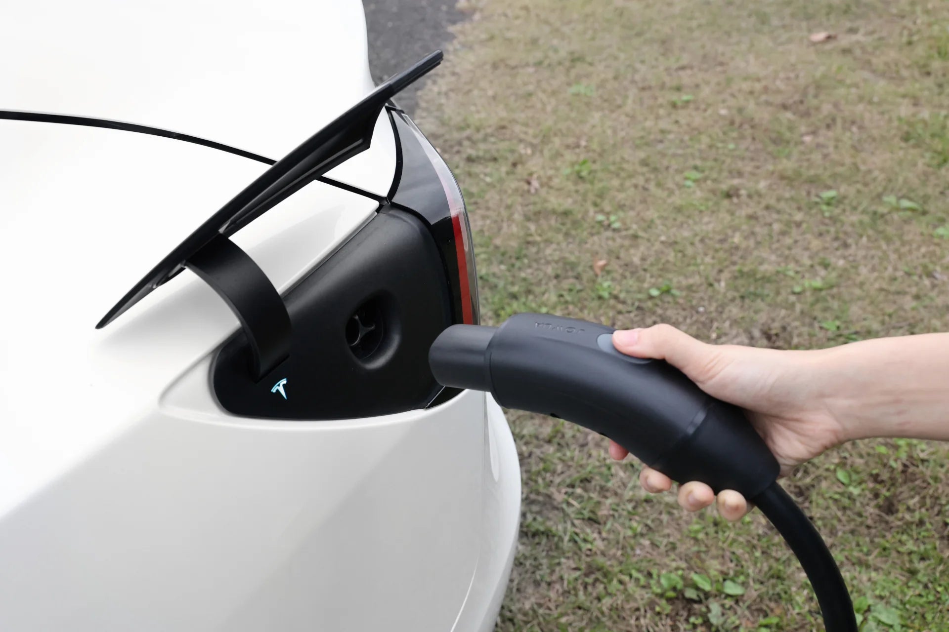 A person plugs a JOWUA NACS Mobile Connector Solution for Tesla (Korea) into the charging port of a white EV with an open flap, parked on grass.