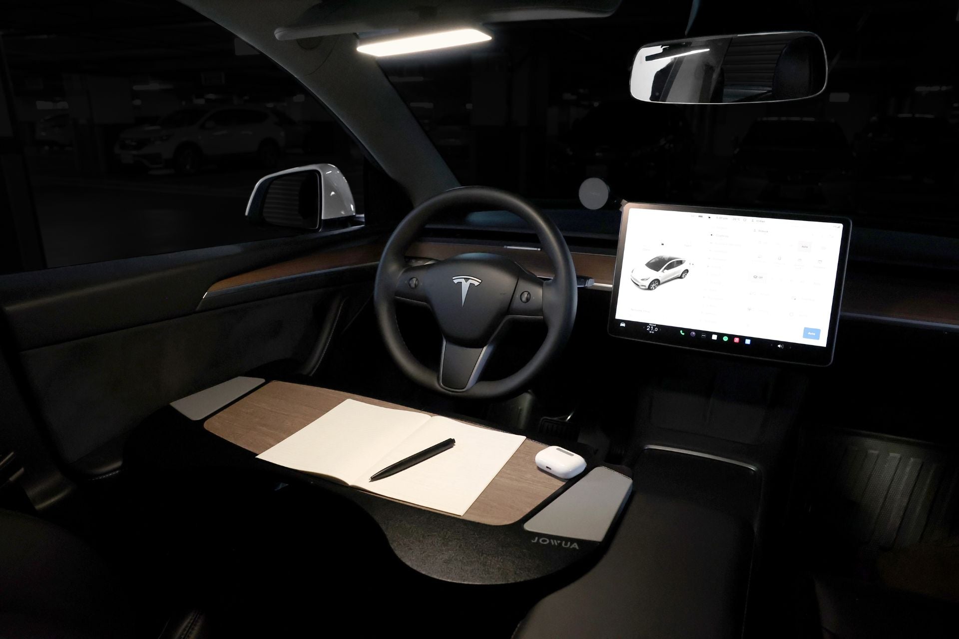 Inside the Tesla car parked in a dimly lit area, the dashboard features a large touchscreen displaying a car interface. Alongside the steering wheel and side mirror, JOWUA