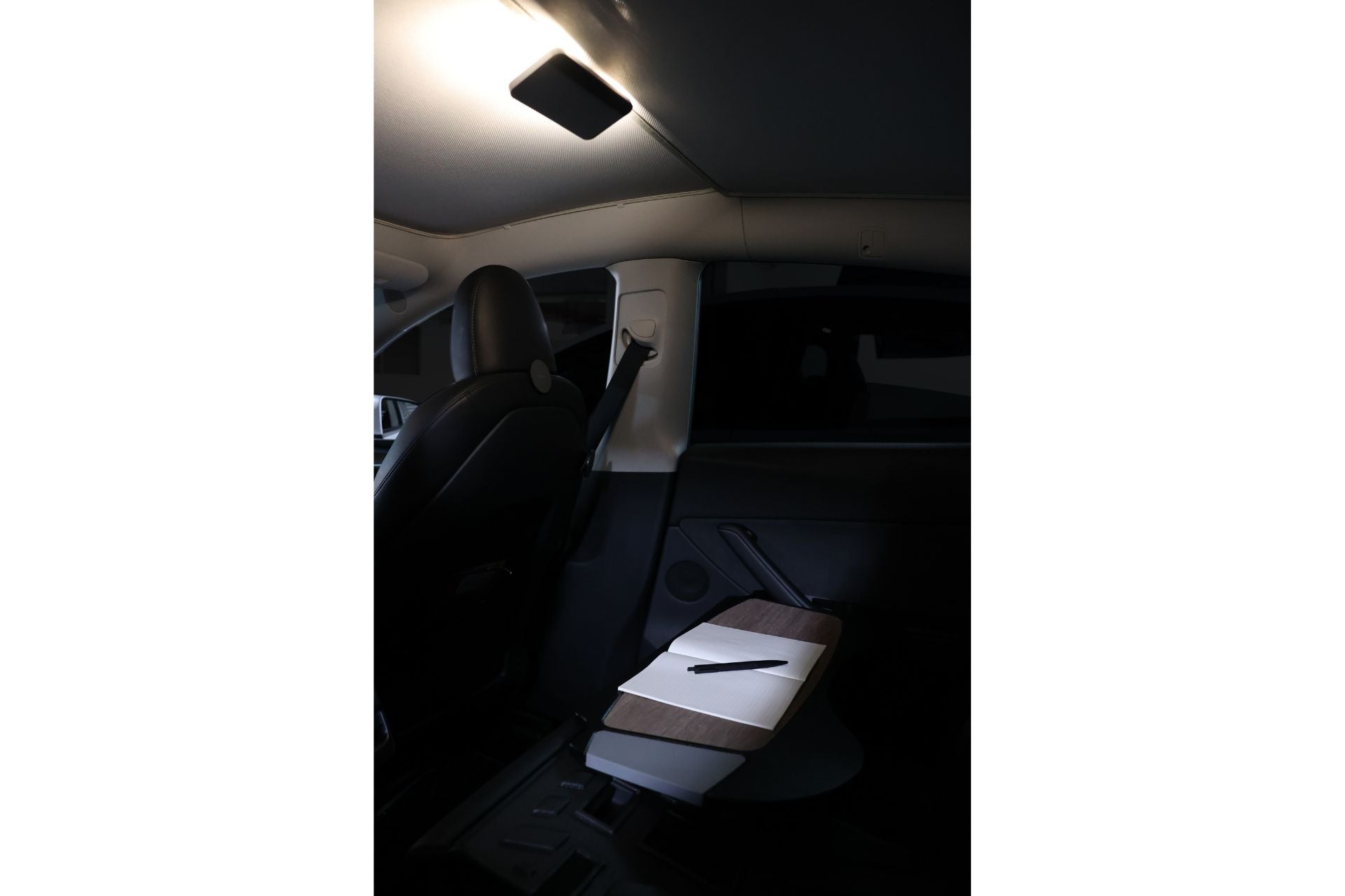 The interior of the car is softly illuminated by a ceiling light, highlighting a wooden folding table with a white surface and a pen on it in the backseat area. To the left, the driver