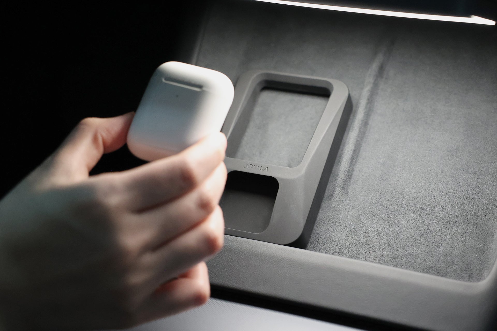 A hand holds a white Apple earbud charging case near the JOWUA Power Dock Combo, an accessory charger for Tesla, resting on a soft surface—possibly inside a car.
