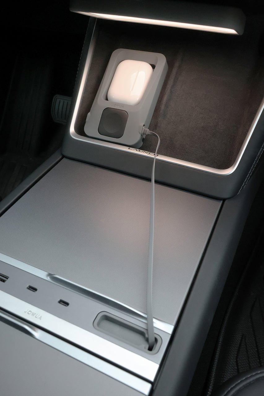 A compact gray electronic device with a glowing white indicator is plugged in and charging on the center console of a car, which boasts a metallic finish and multiple control buttons. The setup features the JOWUA Power Dock Combo – an accessory charger designed for Tesla vehicles – with a discreet MagSafe magnetic charger ensuring reliable power delivery.
