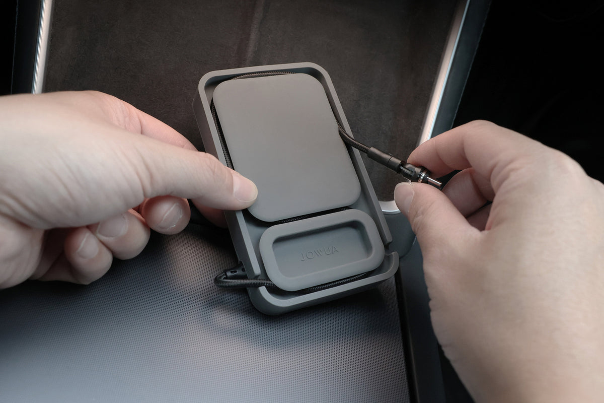 A person holds the JOWUA Power Dock Combo accessory charger with one hand and plugs in a black cable with the other hand inside a Tesla, using it as a charging dock for AirPods.