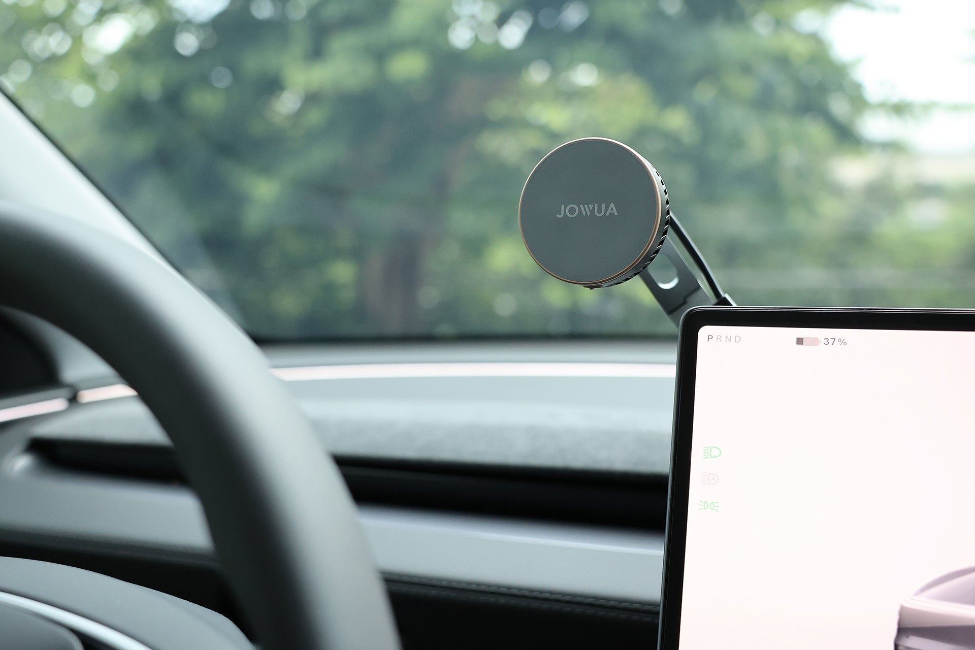 A JOWUA Active Cooling Tesla Compatible Phone Mount (S/3/X/Y/Cybertruck) is attached to a car’s dashboard near a digital display, with the steering wheel and blurred green trees visible in the background.