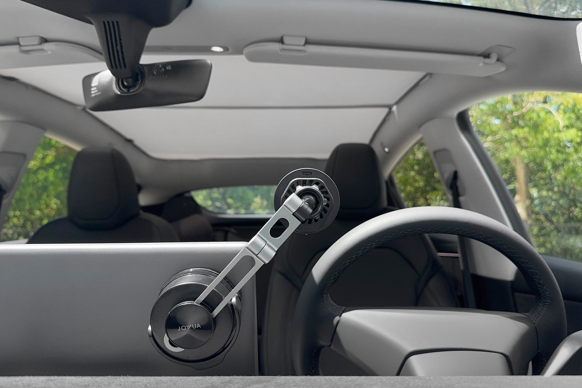 View from the back seat of a Tesla shows a sleek interior with seats, steering wheel, and a JOWUA Active Cooling 6D Foldaway Car Mount Qi2 Wireless Charger (MagSafe) on the dashboard. Lush green trees are visible through the windshield.