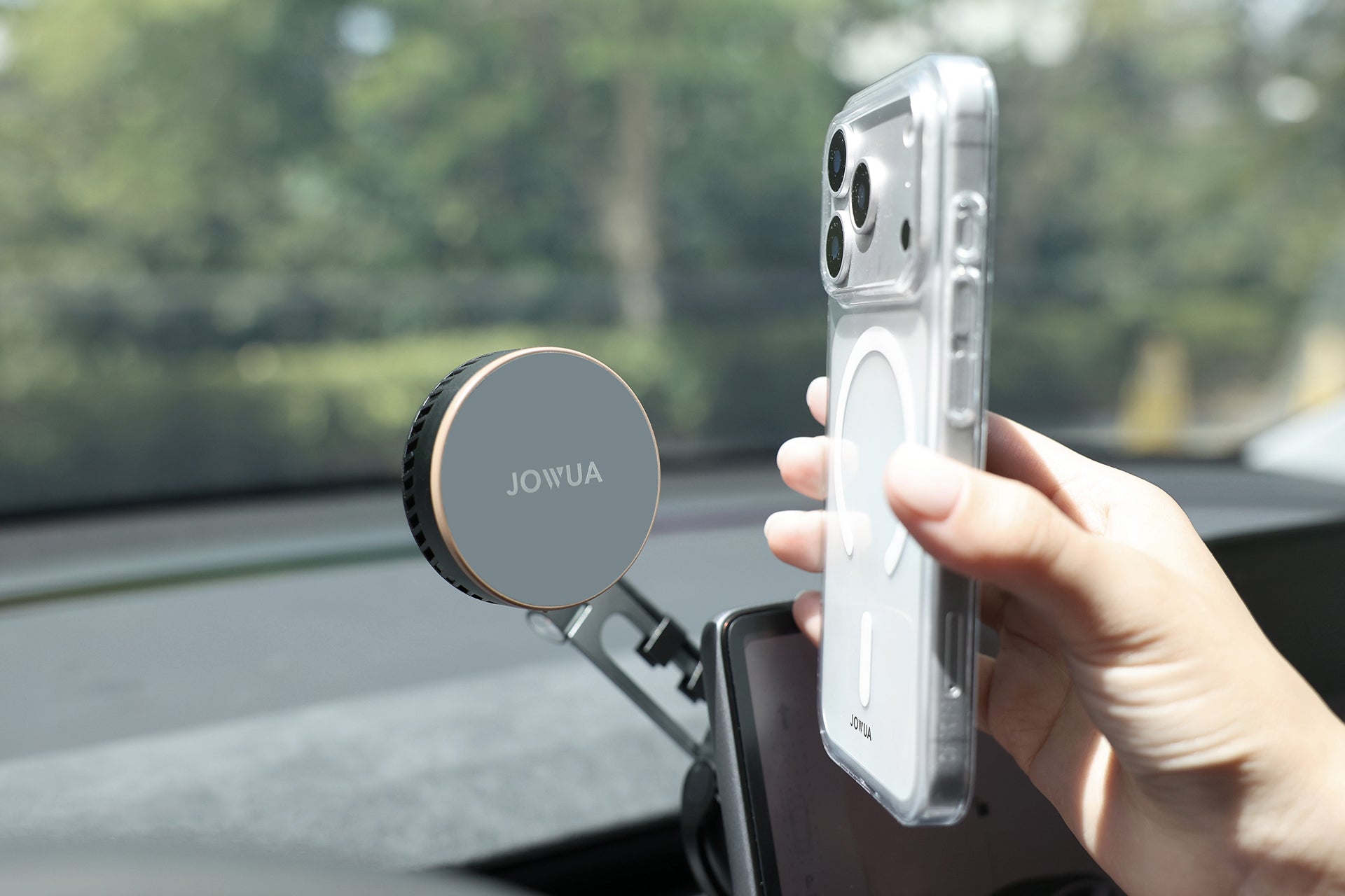 A hand holds a smartphone with a clear case near a JOWUA Active Cooling Tesla Compatible Phone Mount (S/3/X/Y/Cybertruck) on a car dashboard, featuring Qi2 wireless charging, with greenery visible through the windshield.