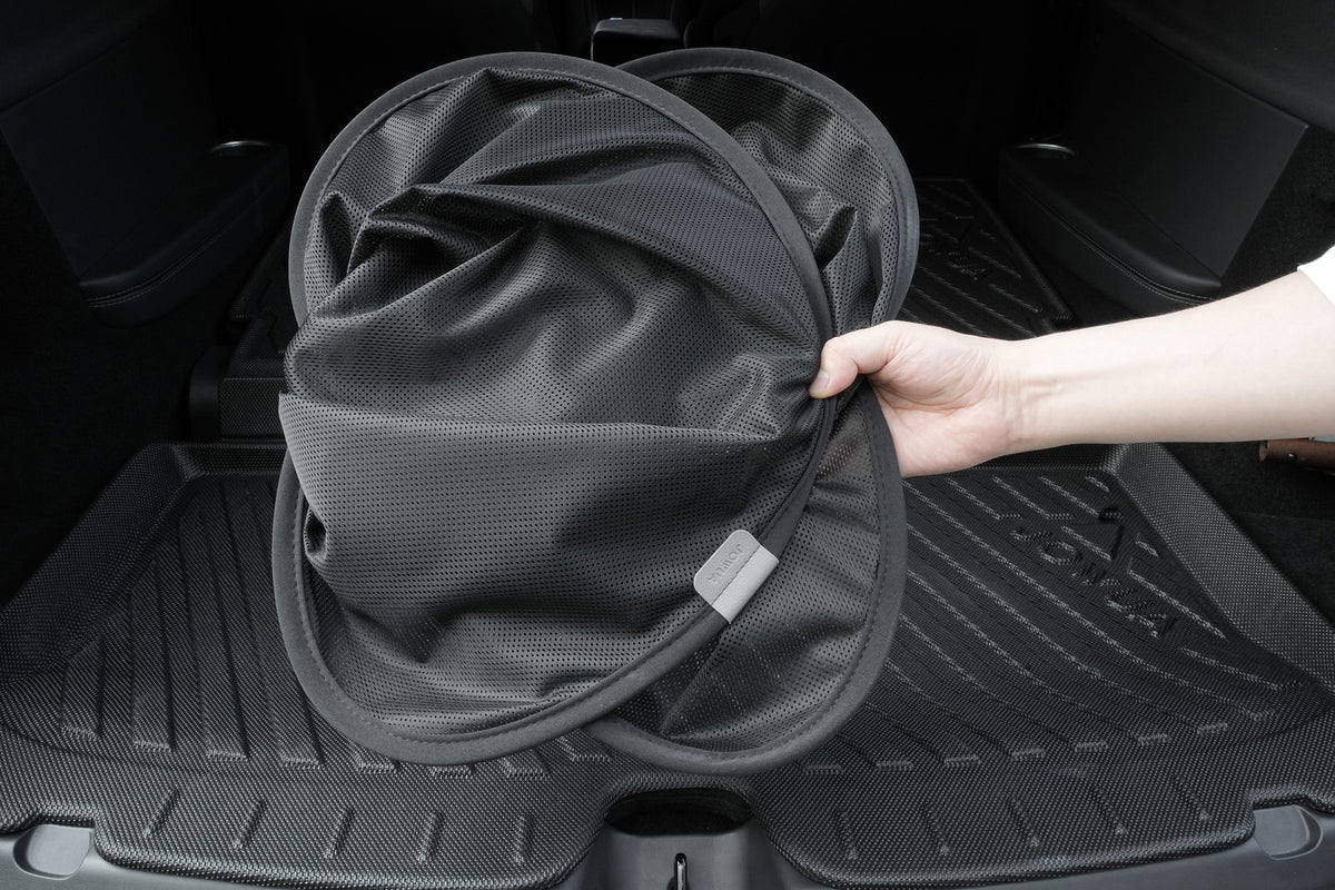 A hand holds a collapsible black mesh storage bin in the trunk of a vehicle with black floor mats, while the JOWUA Rear Lift Gate + Triangular Window Sunshade for Model Y L (2026+) ensures the interior stays cool and protected.