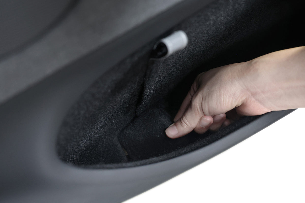 A hand reaches into a black, carpeted compartment to access the JOWUA Reflective Rear Door Emergency Release Strap for Model 3 Highland (2024+) and all Model Y vehicles.