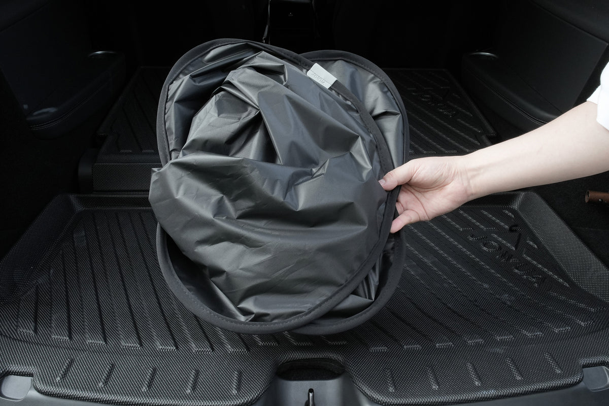 A hand is holding a collapsed, circular black storage bin in the trunk of a vehicle with a textured rubber mat, next to the JOWUA Side + Rear Lift Gate Window Sunshade for Model Y L (2026+) for added organization and protection.