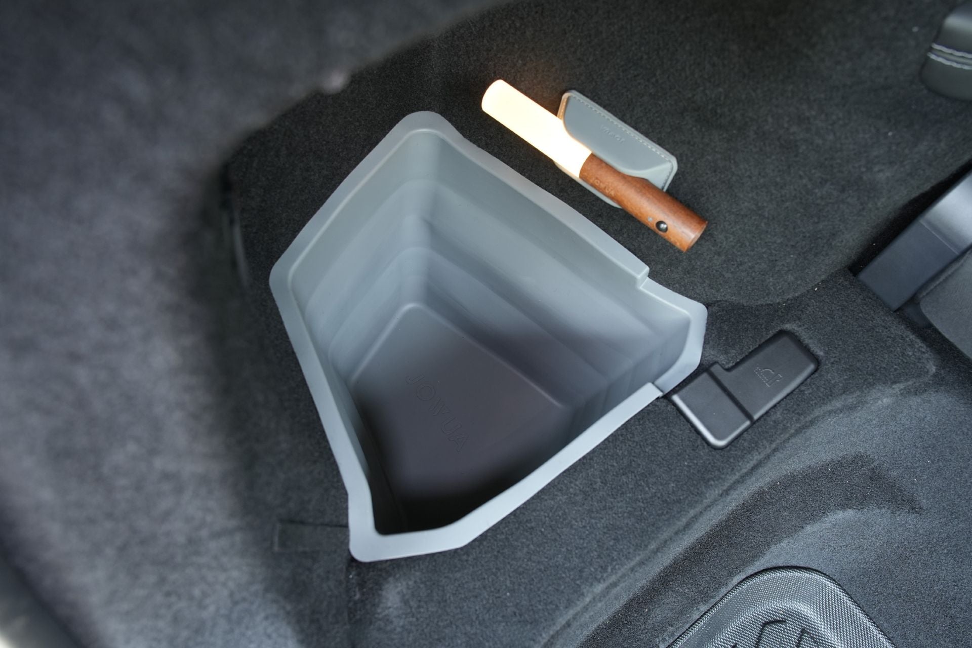 A gray, shallow JOWUA Tesla Model Y L (2026+) Trunk Storage Bin from the Ambient Light Bundle is integrated into the trunk floor, positioned beside a wooden-handled flashlight in its holder, and surrounded by dark carpeting.