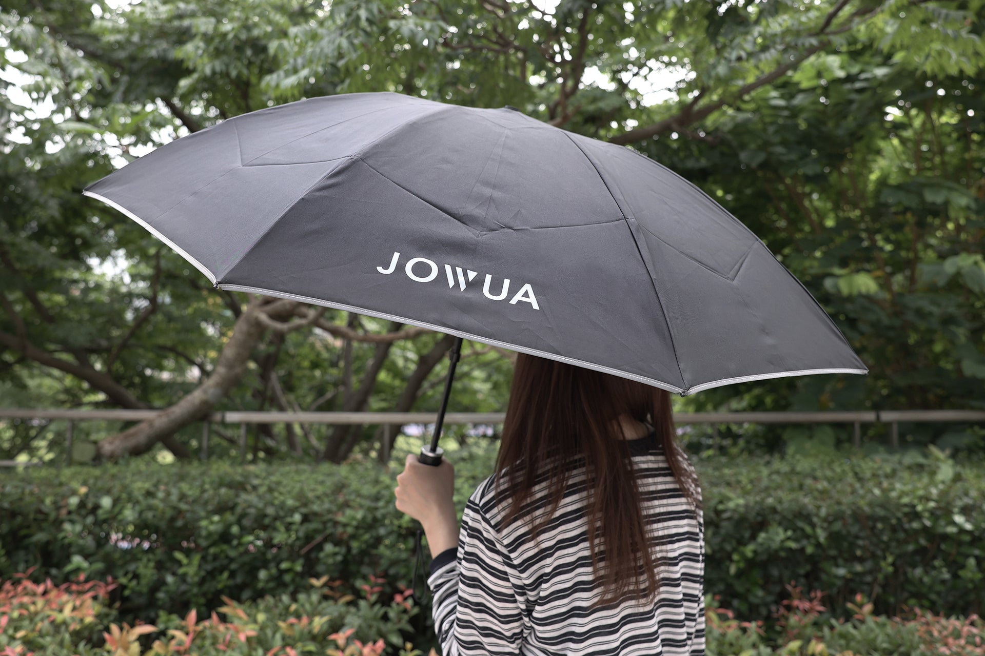 A person with long hair in a striped shirt holds a JOWUA Type-C Electric Umbrella, featuring USB Type-C charging, outdoors near trees and greenery.