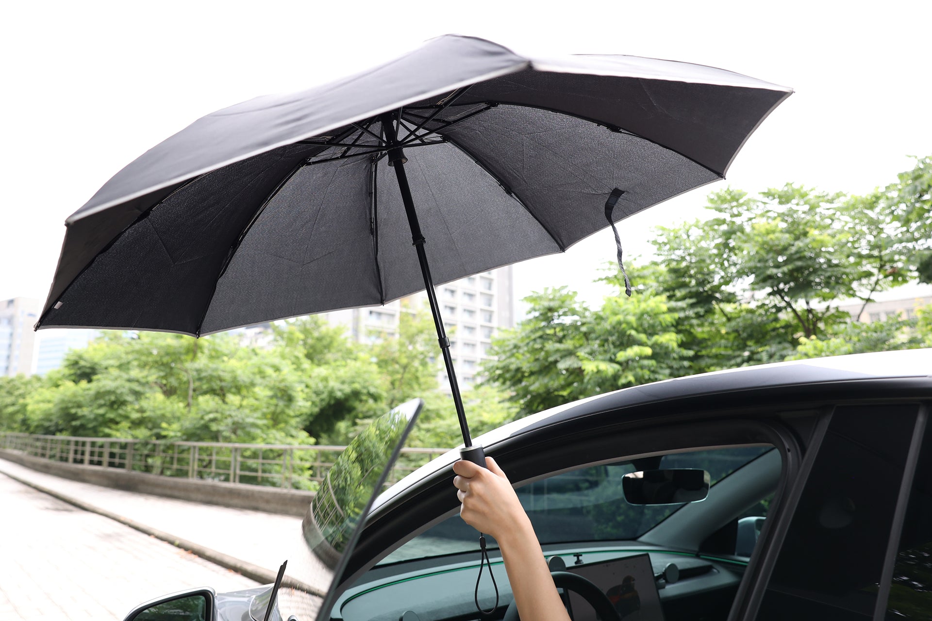A person’s hand holds a JOWUA Type-C Electric Umbrella with reverse folding outside a car door, with green trees and buildings in the background.