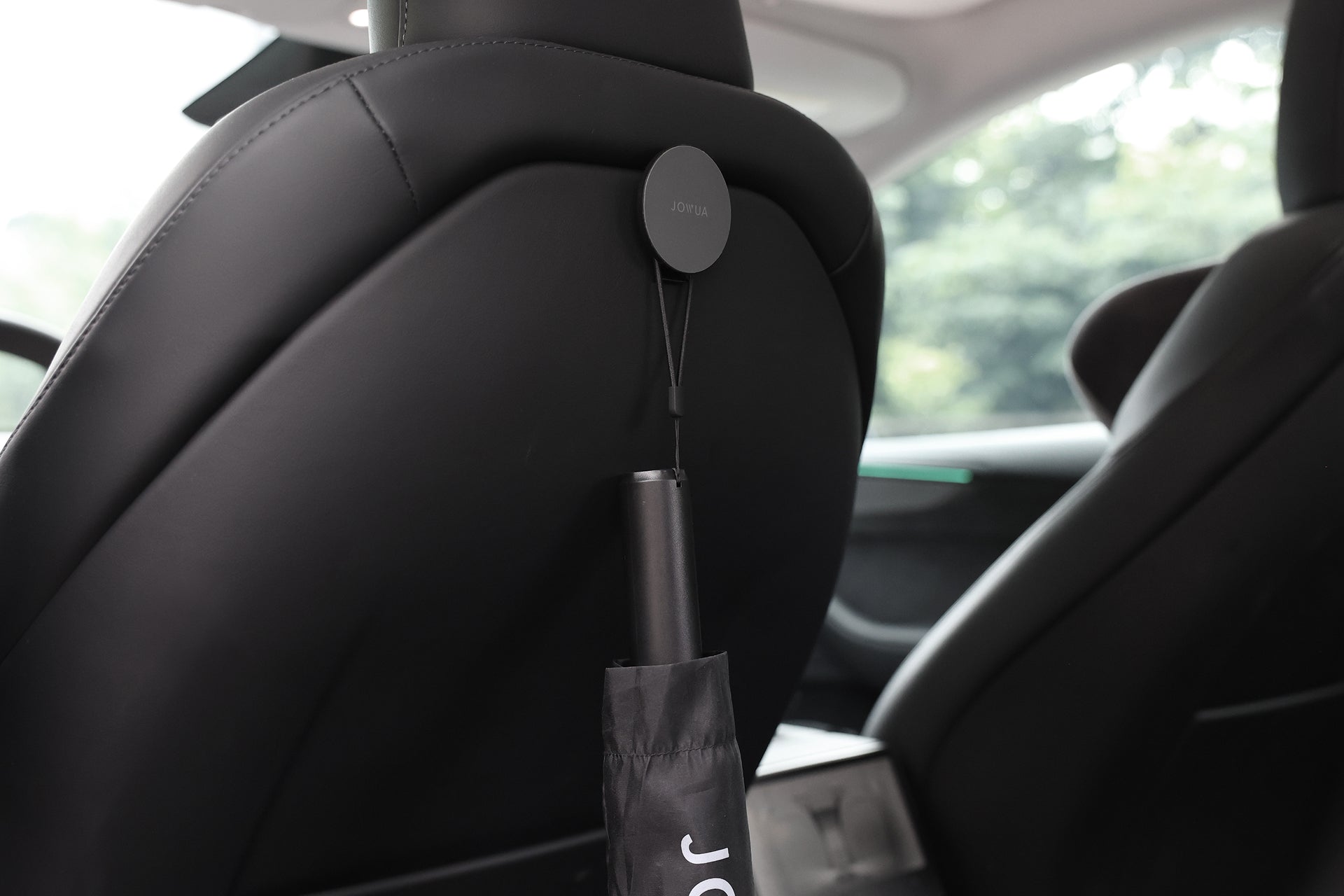 A JOWUA Type-C Electric Umbrella in its case hangs from a car seat headrest inside a modern vehicle, with green trees blurred outside the window.