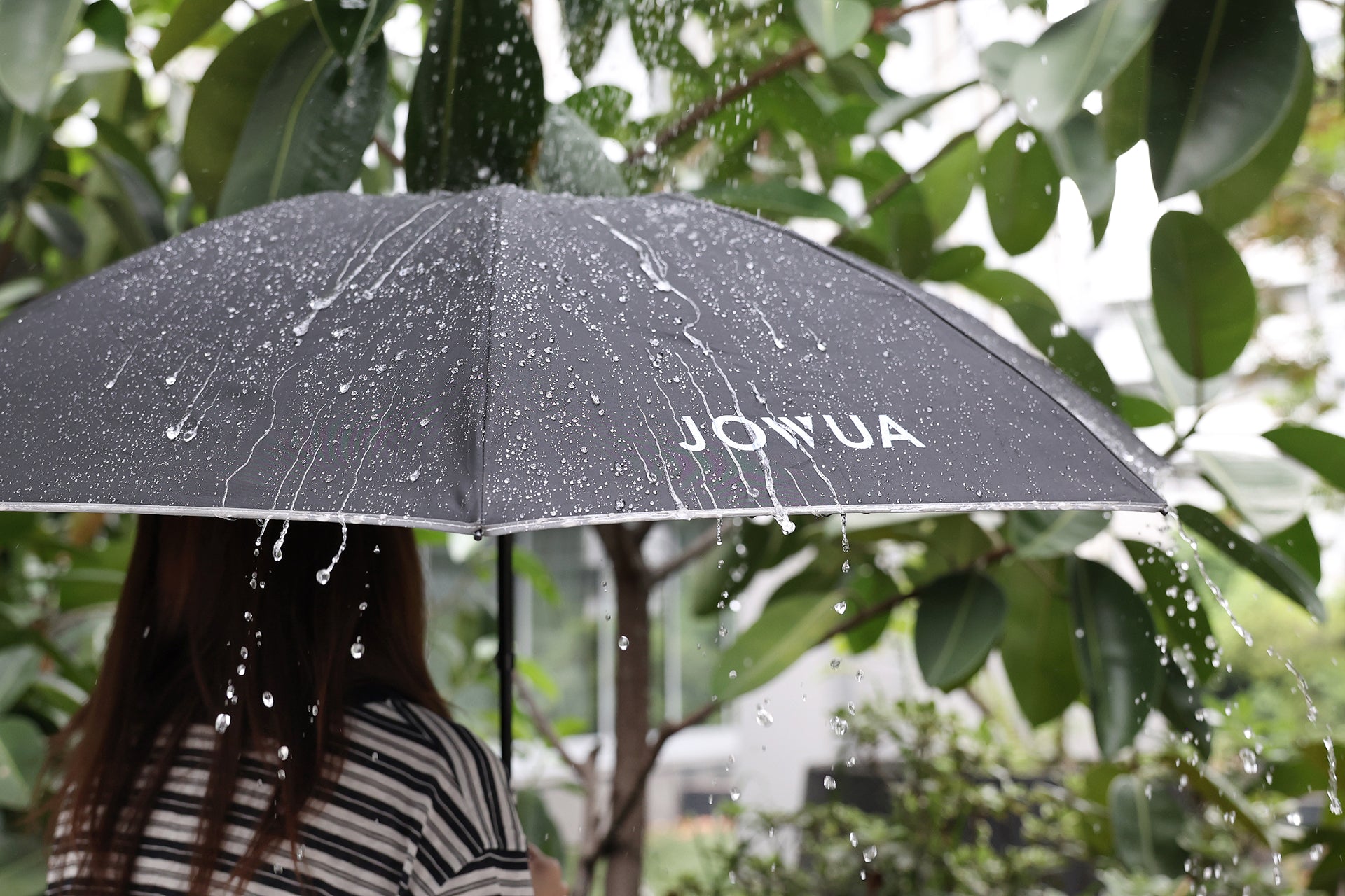 A person stands in the rain among green plants, holding a black JOWUA Type-C Electric Umbrella with visible water droplets on its reverse folding canopy.