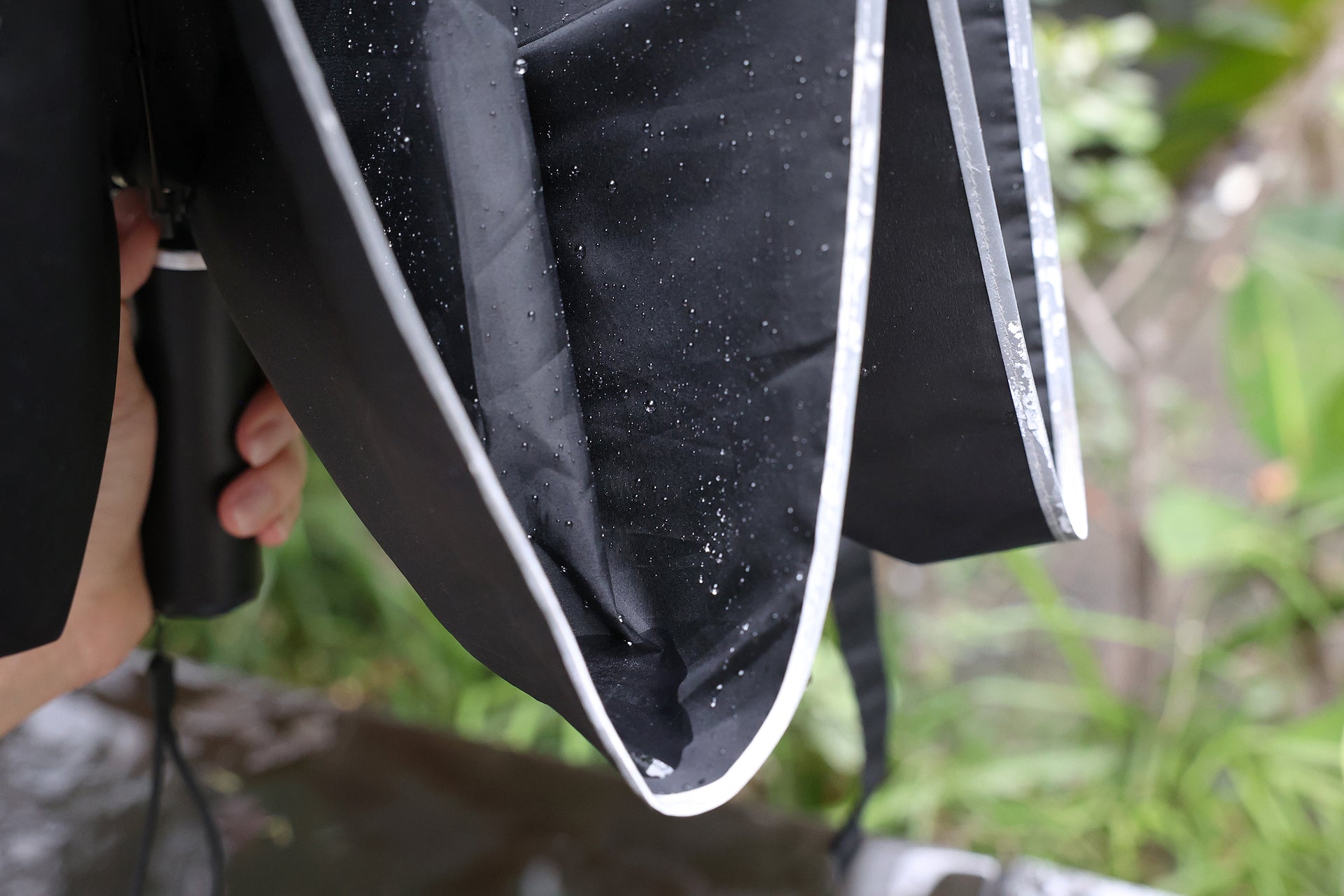 A close-up shows a person holding the JOWUA Type-C Electric Umbrella, featuring a wet black fabric with white trim and glistening water droplets, while lush greenery forms a blurred background.