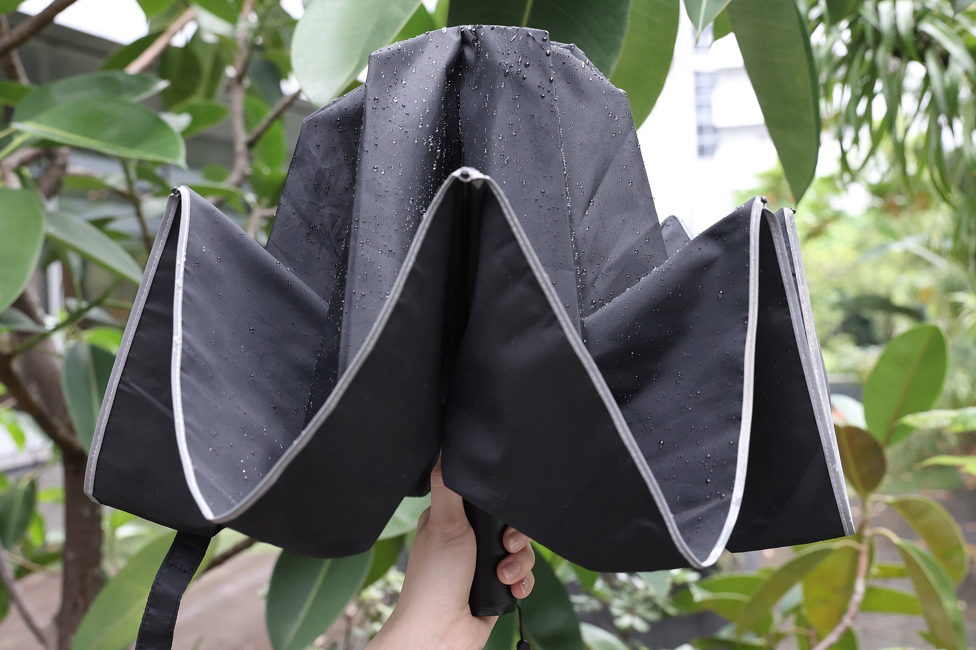 A hand holds a JOWUA Type-C Electric Umbrella, shown upside-down with raindrops on its black surface, surrounded by green plants and trees outdoors.