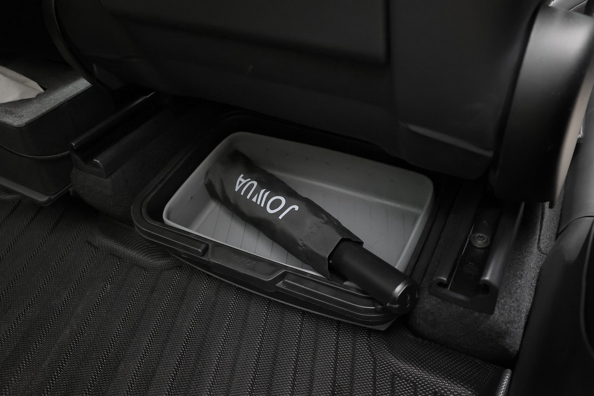 A JOWUA Type-C Electric Umbrella with white text lies in a plastic tray under a car seat, placed on a black textured floor mat.