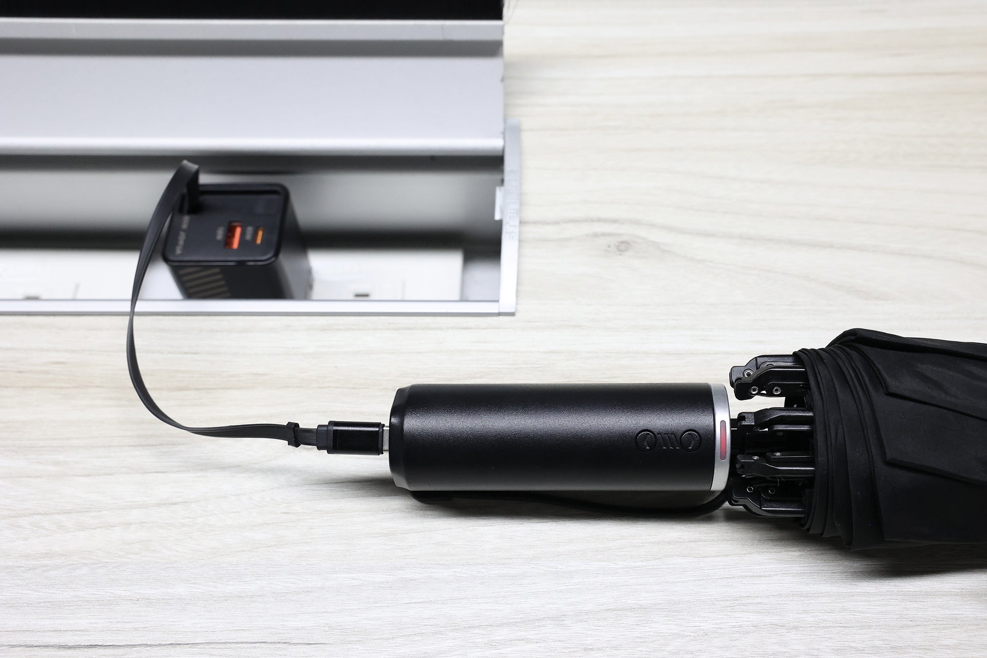A JOWUA Type-C Electric Umbrella with a built-in handle is plugged into a USB Type-C port on a light wooden desk beside an open power strip.