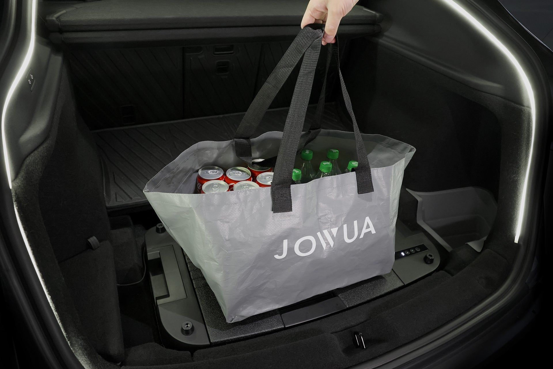 A hand lifts a gray JOWUA Car Refrigerator for Tesla Model Y filled with soda cans and green bottles from a trunk—ideal for road trips with an intelligent dual-mode cooling system to keep your drinks chilled.