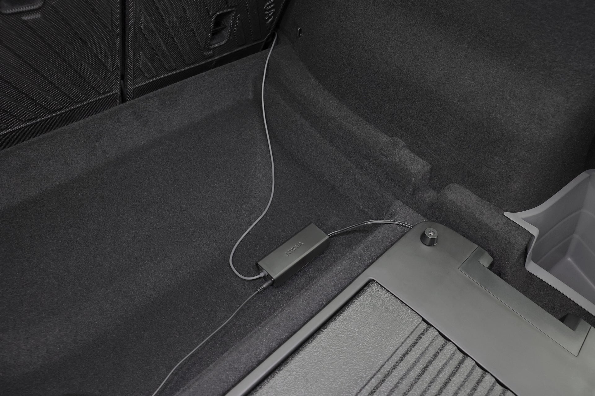 A close-up of a Tesla Model Y trunk shows a JOWUA Car Refrigerator with dark carpeting and a charging cable connected, lying next to a plastic storage compartment.