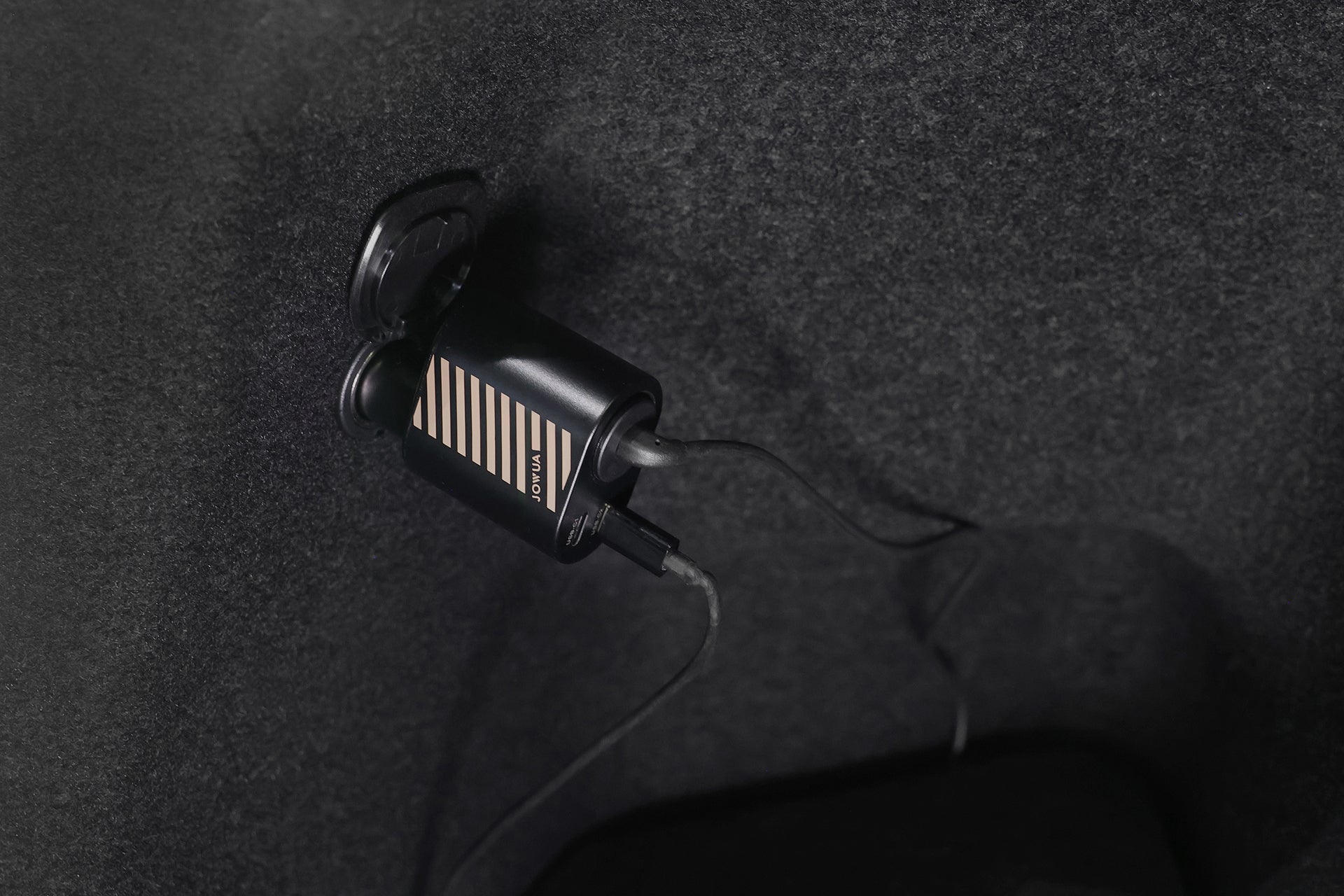 A close-up of the JOWUA Car Refrigerator for Tesla Model Y plugged into a wall socket with USB-C power mode, showing its connected cable extending down against a dark, textured background.