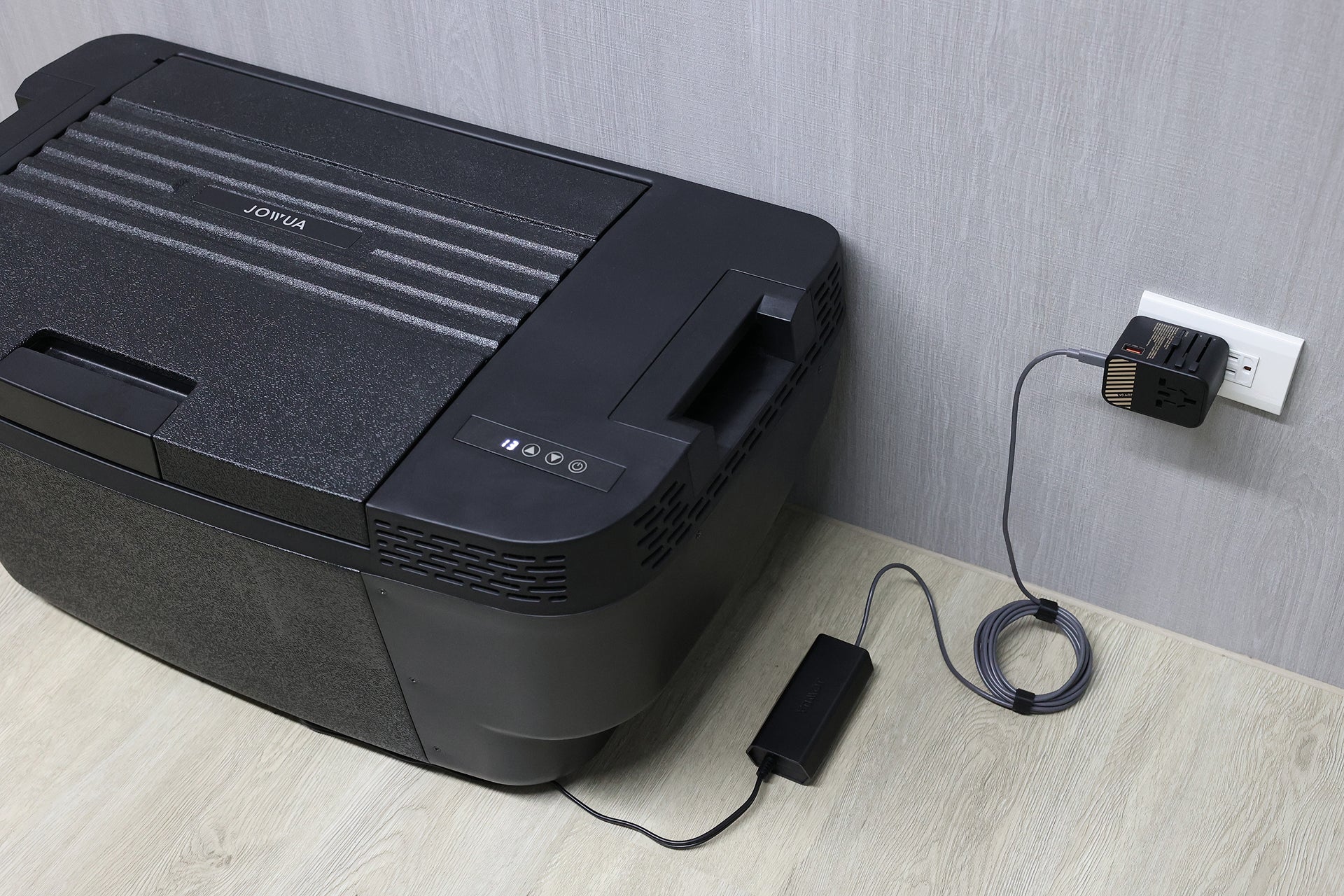 The JOWUA Car Refrigerator for Tesla Model Y, in black, features intelligent dual-mode cooling and is powered by USB-C with an adapter and coiled cord. It sits on a light wood floor next to a gray wall.