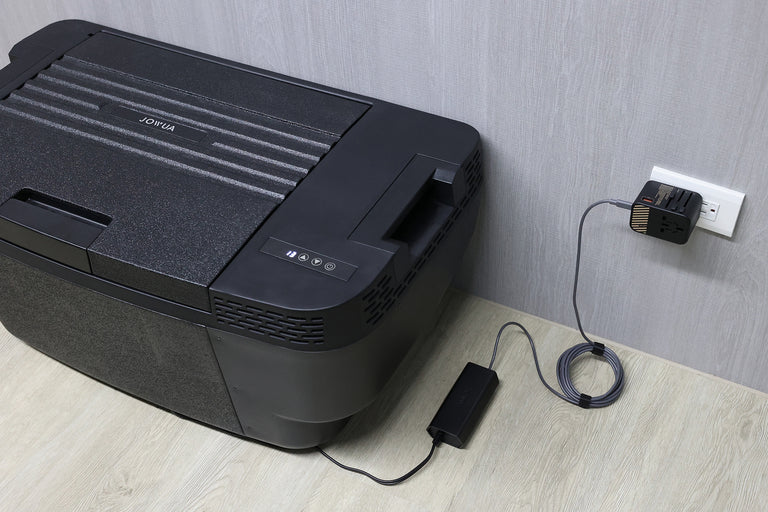 The JOWUA Car Refrigerator for Tesla Model Y, in black with intelligent dual-mode cooling, is plugged into a wall outlet by a power adapter and cable, and placed on a light wood floor beside a gray wall.