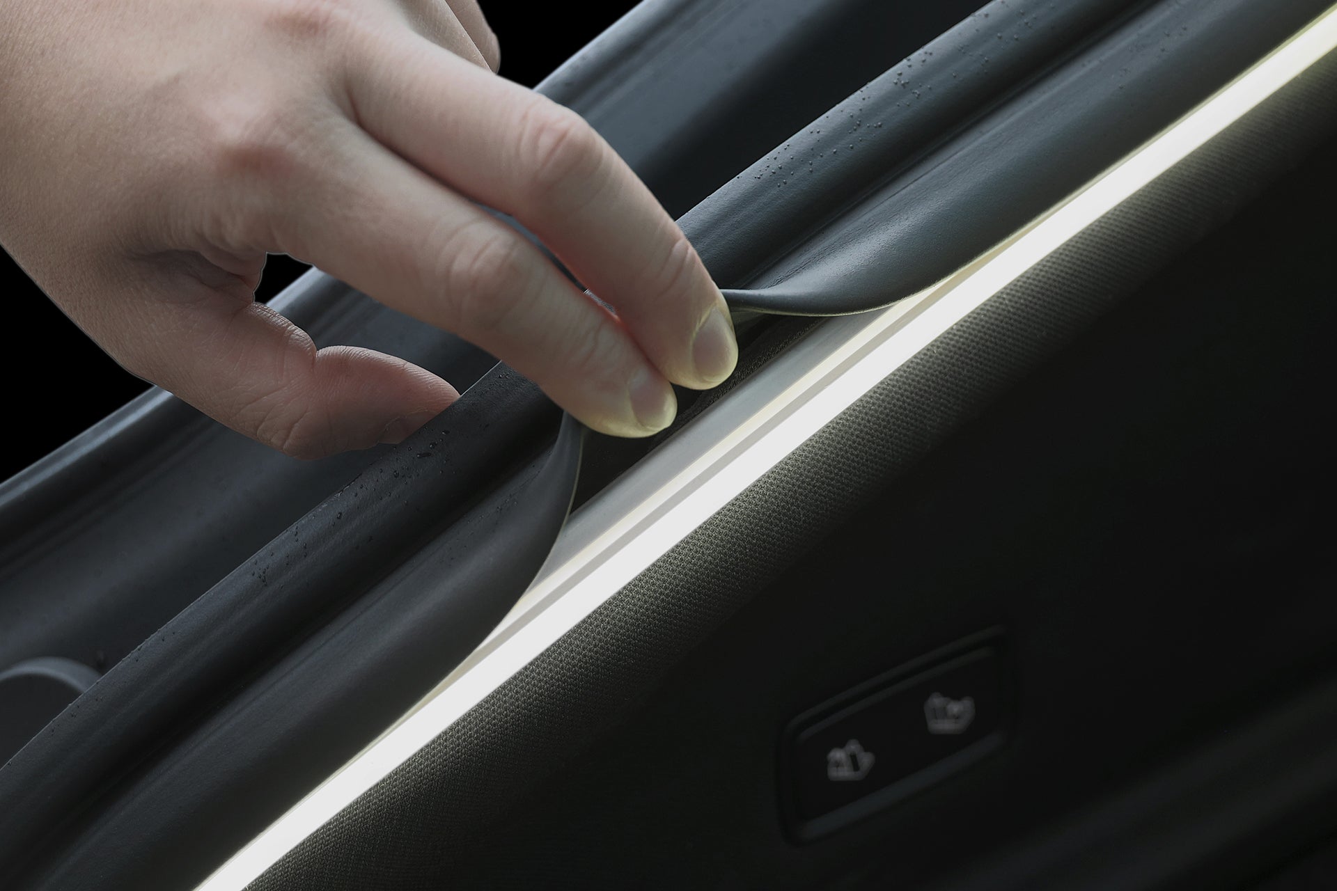 A close-up of a hand lifting a rubber car door seal to reveal the JOWUA Tesla Model Y Trunk Ambient Light, a USB-C LED strip with magnetic induction auto-switch, installed beside the window control button.