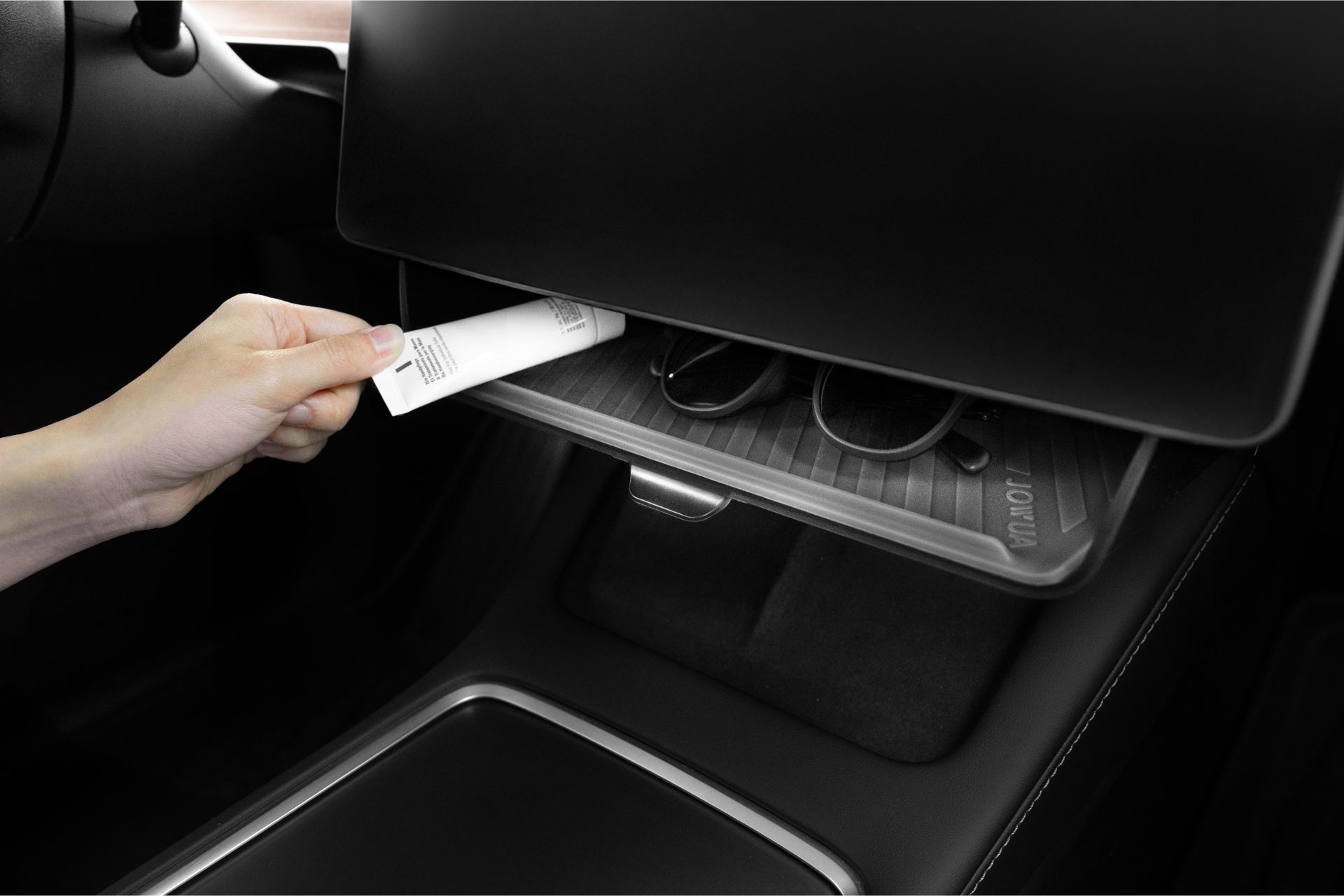 A hand places a small tube into a Tesla's dashboard tray next to glasses, highlighting JOWUA’s Under Screen Invisible Station for Model 3 Highland (2024+)/Y Juniper (2025+), designed for minimalist style and seamless accessory integration.