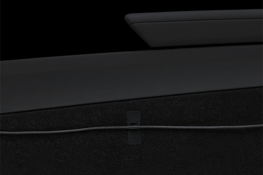 Close-up of a black interior car panel with a sleek armrest above a dark carpeted surface. A JOWUA Magnetic Charging Cable USB-C to USB-C (1M-1.8M) runs horizontally across the carpet, secured in place by a small strap—a subtle nod to Tesla Model 3 accessories.