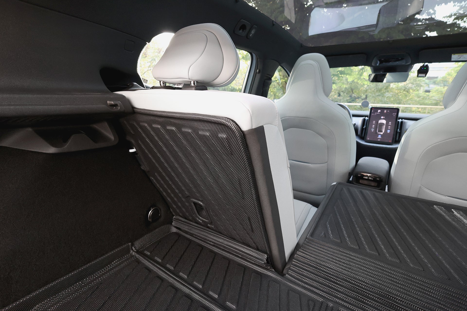 Rear cargo view of a Volvo EX30 with one seat folded, featuring the JOWUA All Weather Trunk Liner + Rear Seat Cover, light gray interior, a large central touchscreen, and trees visible through the windows.