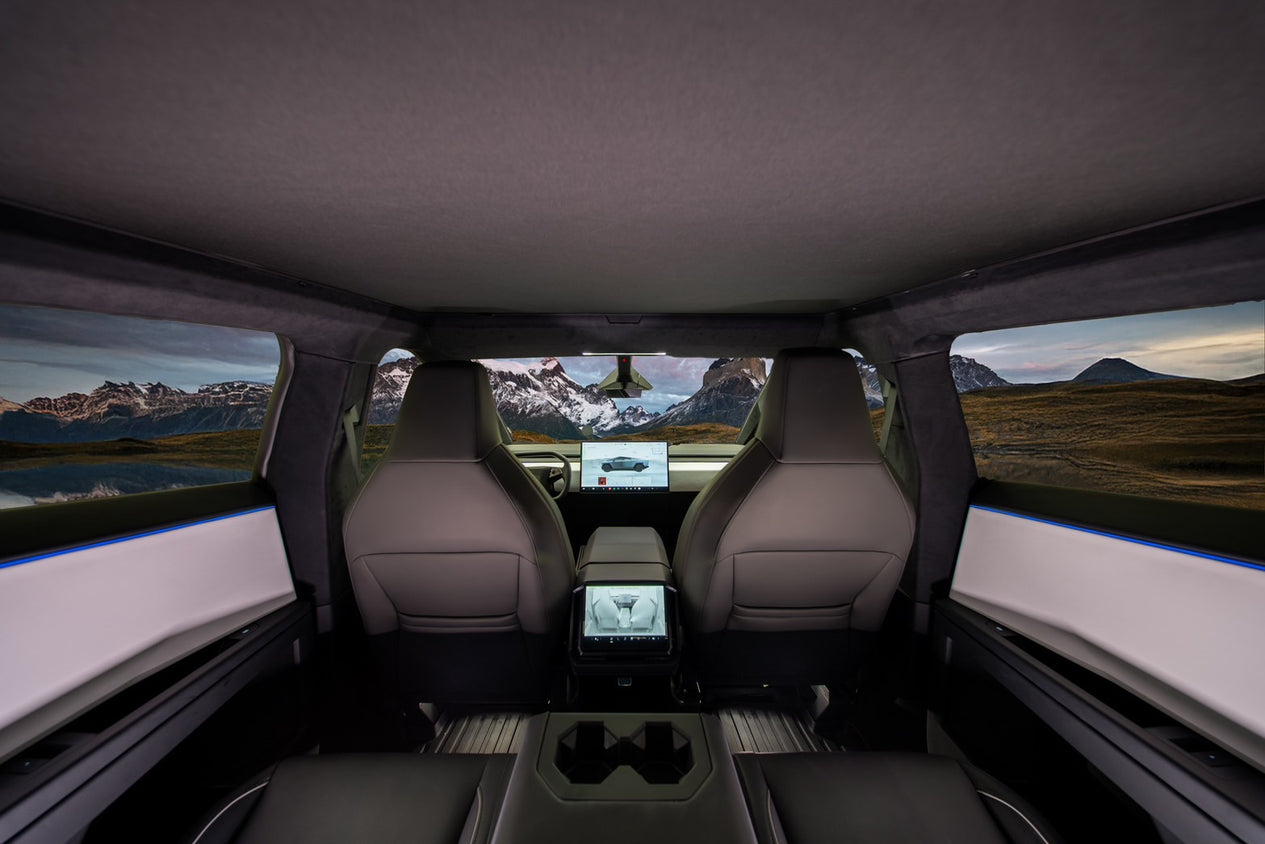 A view from inside a Cybertruck showcases the lightweight mesh fabric sunshade on the two front seats and a dashboard adorned with digital displays. The expansive windows, featuring JOWUA