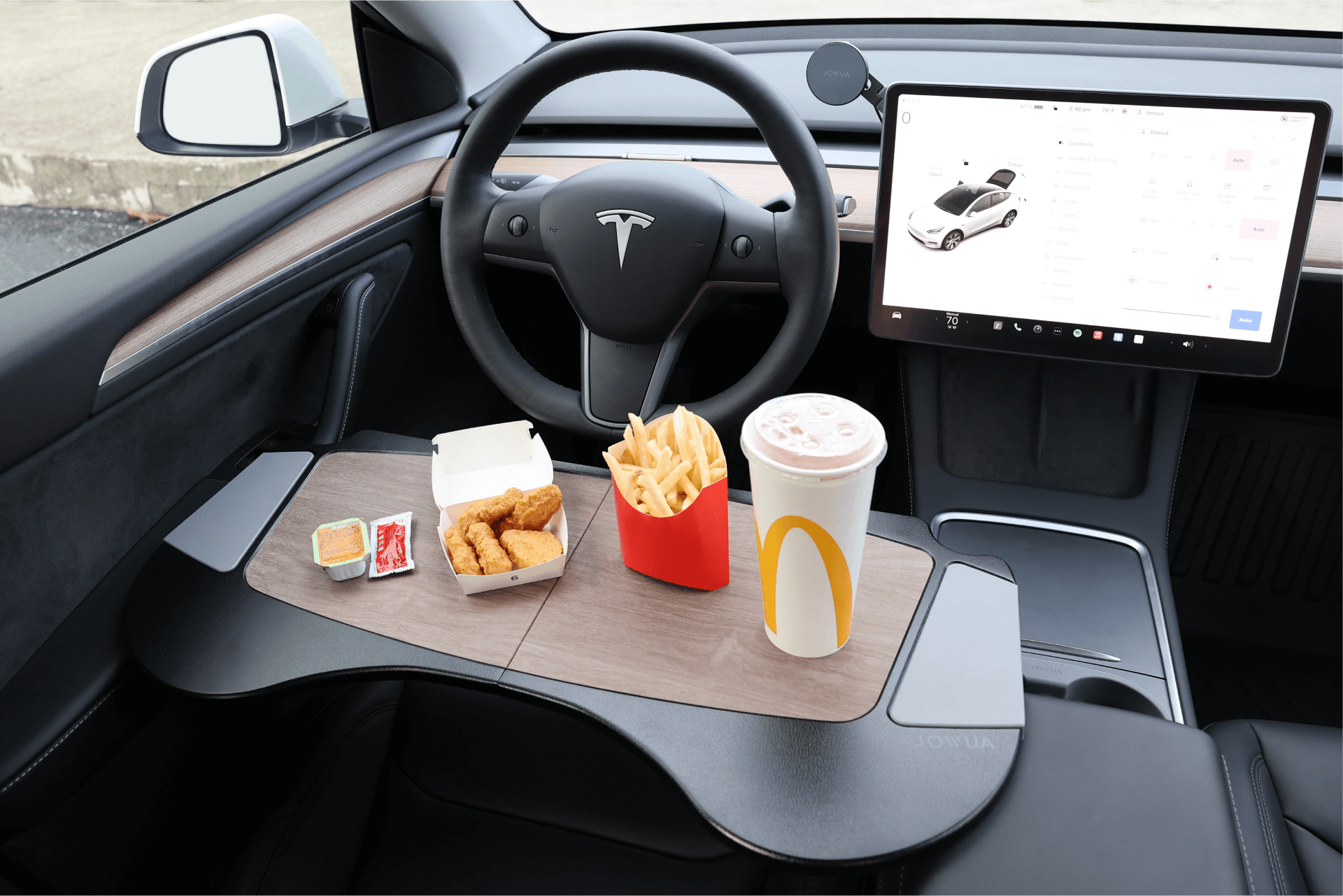 A Foldable Car Tray Bundle by JOWUA is neatly arranged on a Tesla's steering wheel, holding a fast-food meal with fries, chicken nuggets and sauce, and a drink. The car's interior and dashboard screen are visible in the background, highlighting this sleek Tesla accessory.