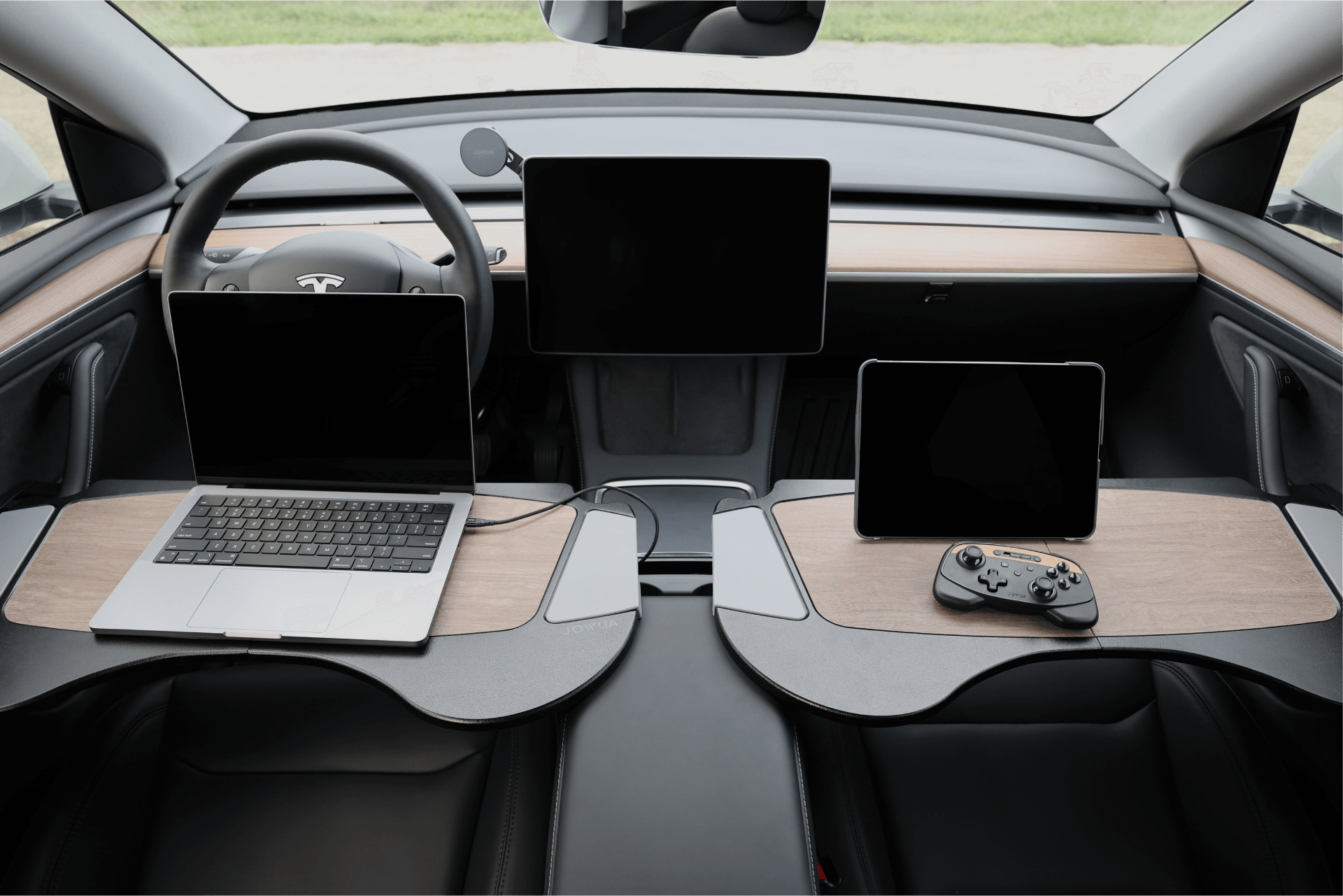 The interior of a Tesla Model S features a steering wheel, central tablet display, and sleek wooden accents. A laptop rests on the left with a handheld gaming console on the right. Enhance your drive with the JOWUA Foldable Car Tray Bundle that complements its minimalist design.