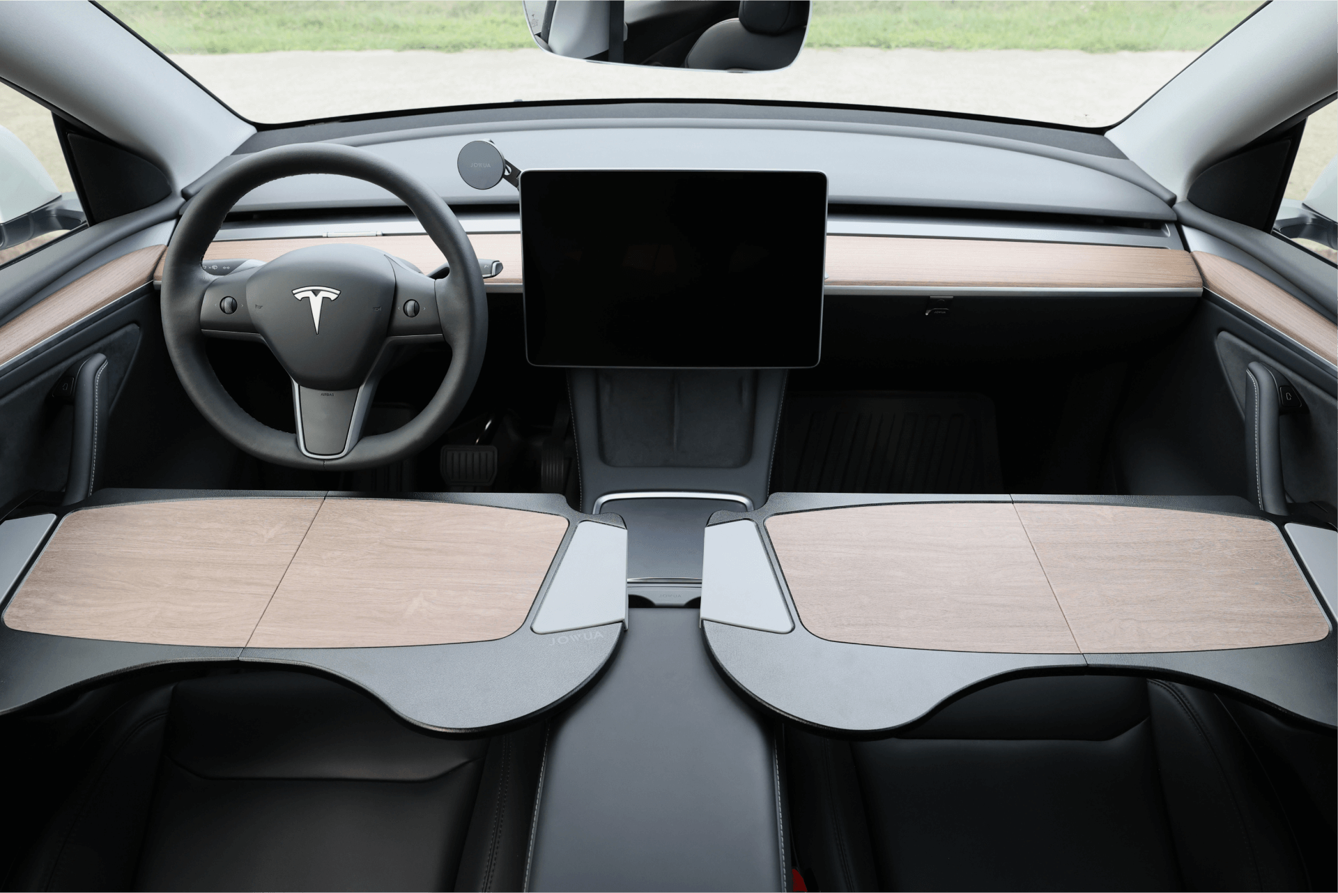 The interior of a Tesla Model Y features a minimalist dashboard with a large central touchscreen and wood-accented panels. Complementing the steering wheel adorned with the Tesla logo is the JOWUA Foldable Car Tray Bundle, which offers convenient functionality. The black seats provide comfort, while the windshield showcases the greenery outside.