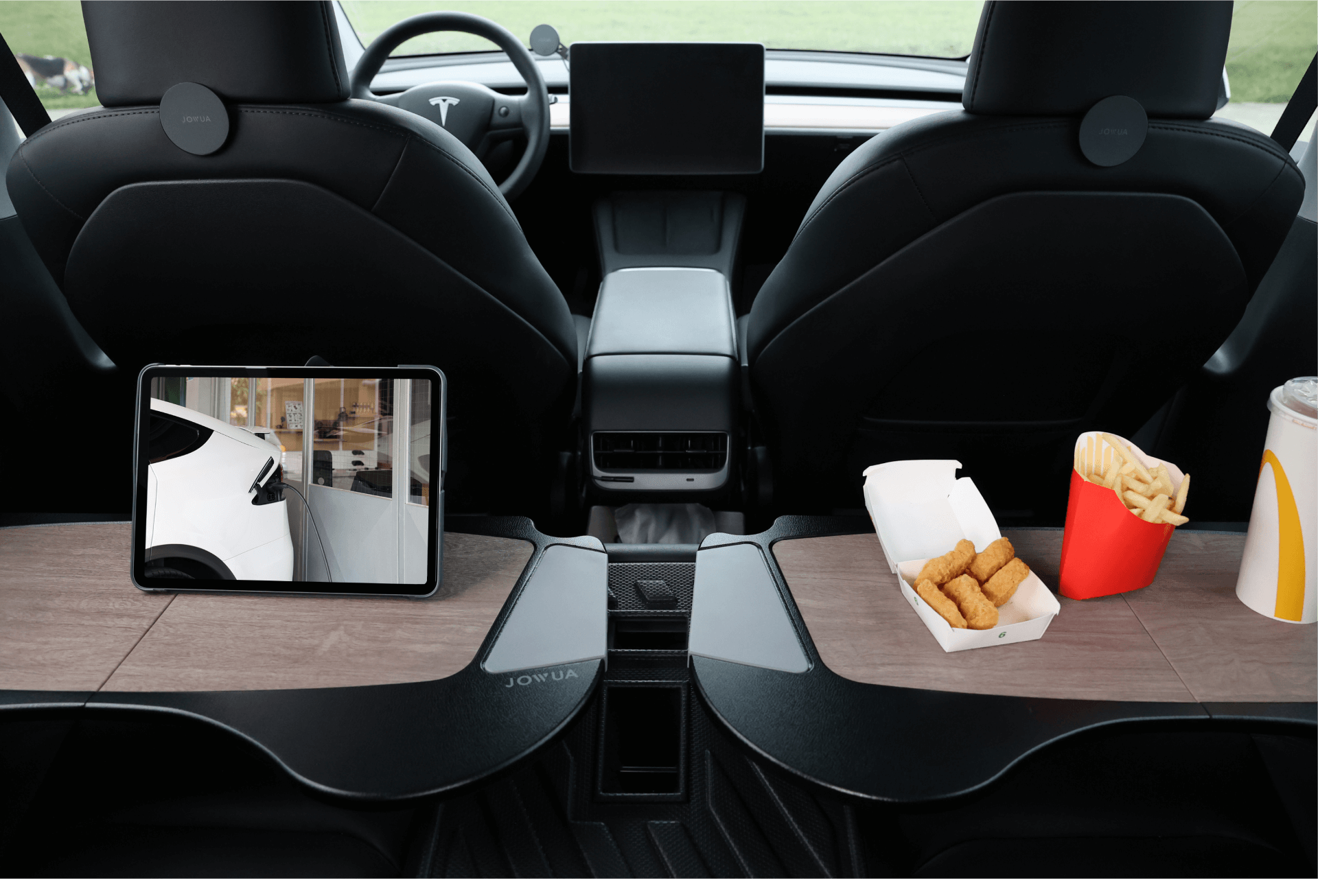 The interior of a Tesla Model Y highlights the JOWUA Foldable Car Tray Bundle: one side supports a tablet displaying an outdoor scene, while the other accommodates fries, nuggets, and a drink with a straw. In the backdrop, you can see the car seats and the distinctive steering wheel.