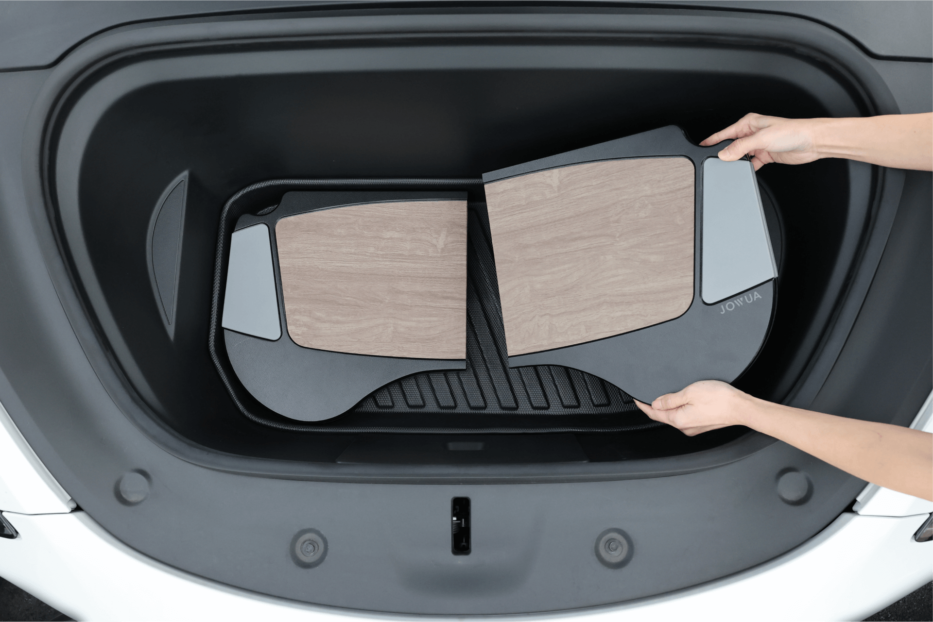 A person places a set of stylish JOWUA Foldable Car Tray Bundles, featuring wood-like panels, into the front storage compartment of a white Tesla. These sleek accessories boast contrasting black and gray elements crafted from durable PU leather, perfectly complementing the vehicle's sophisticated interior.
