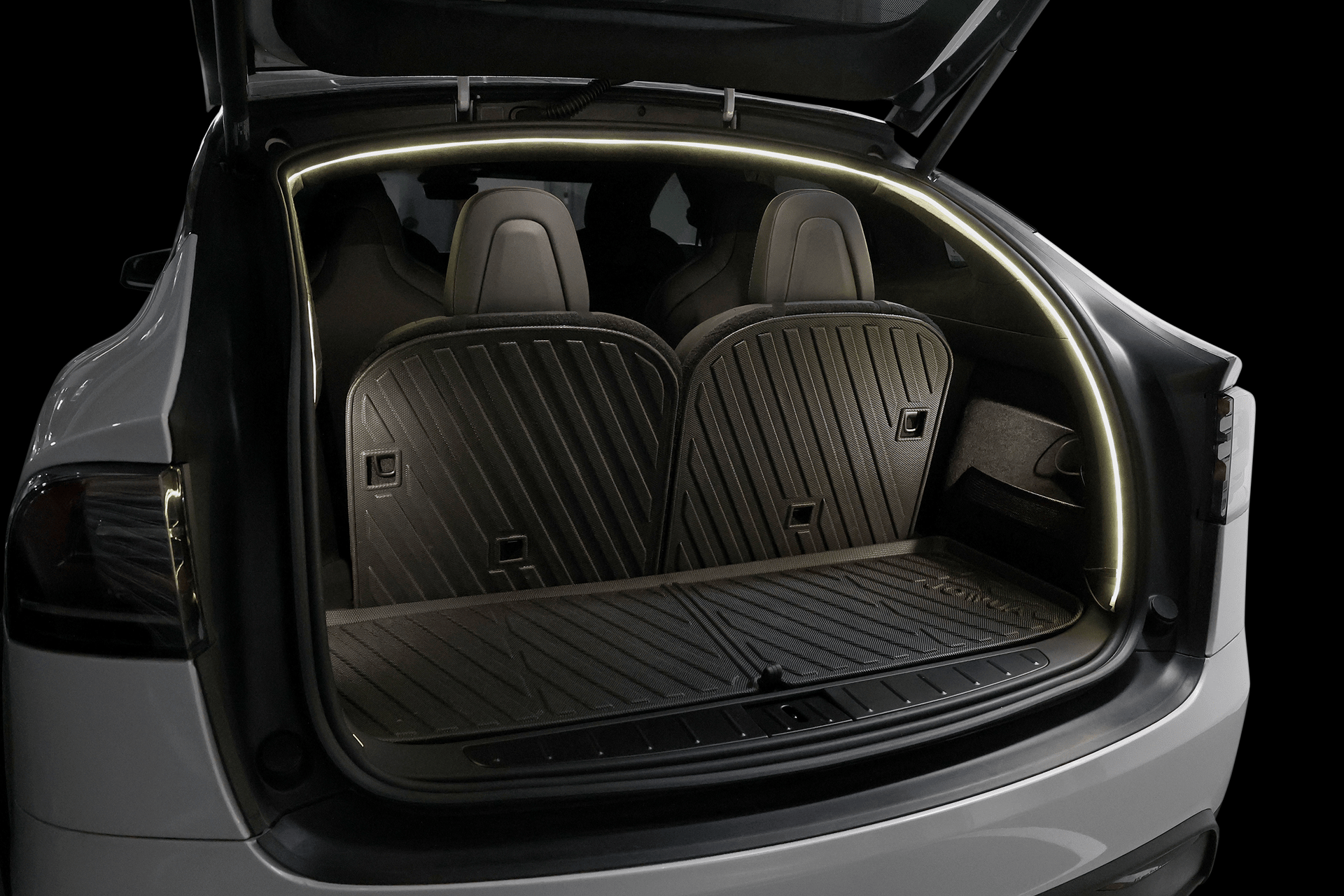 The open trunk of a white Tesla Model X showcases JOWUA’s Trunk Ambient Light - USB-C LED Strip outlining the opening, along with fitted rubber floor mats and visible rear seats in the background.