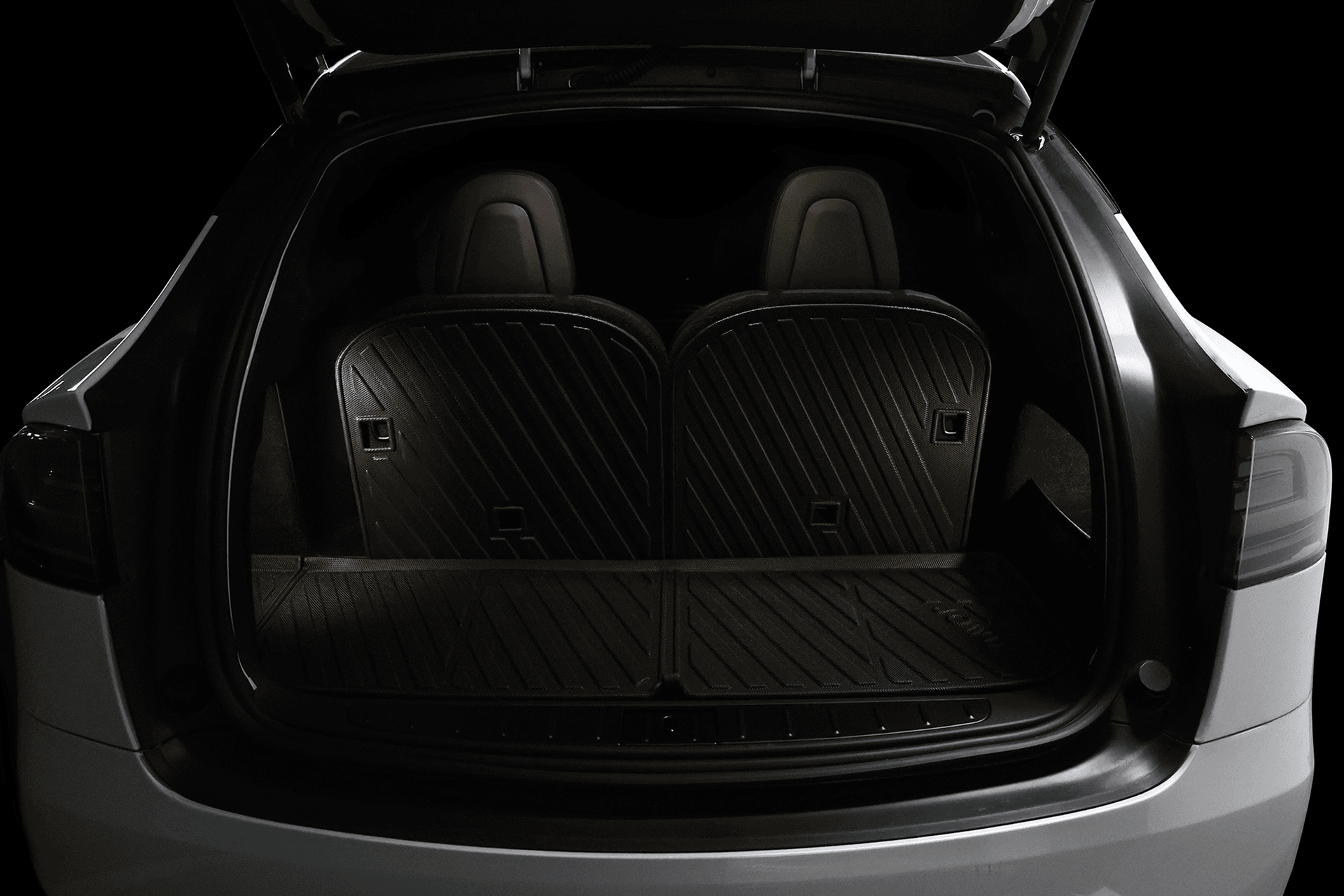 The JOWUA Tesla Model X Trunk Ambient Light - USB-C LED Strip softly illuminates the rear trunk, showcasing black rubber floor mats and two upright rear seats, as seen from the back in low lighting.